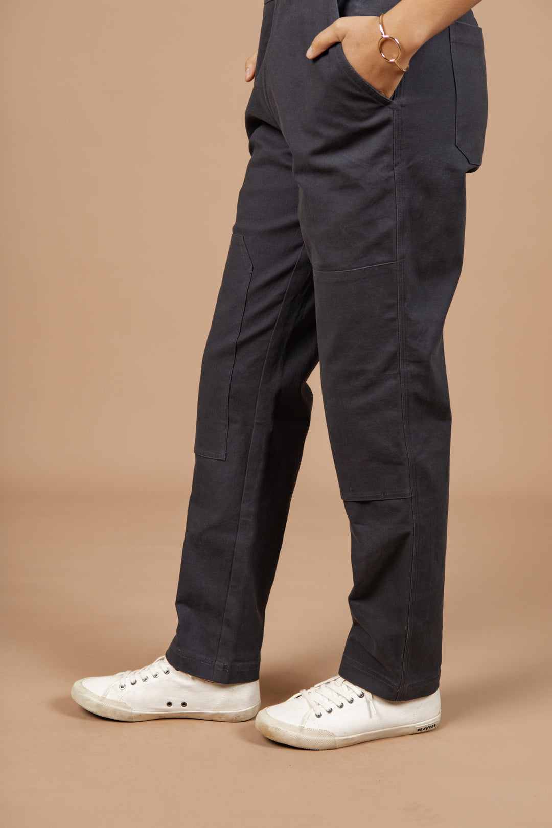 A person stands with hands in pockets, wearing Bridge & Burn's Tobin Utility Overall in Dark Charcoal Canvas and white sneakers. Against a beige background, their upper body is mostly out of frame.