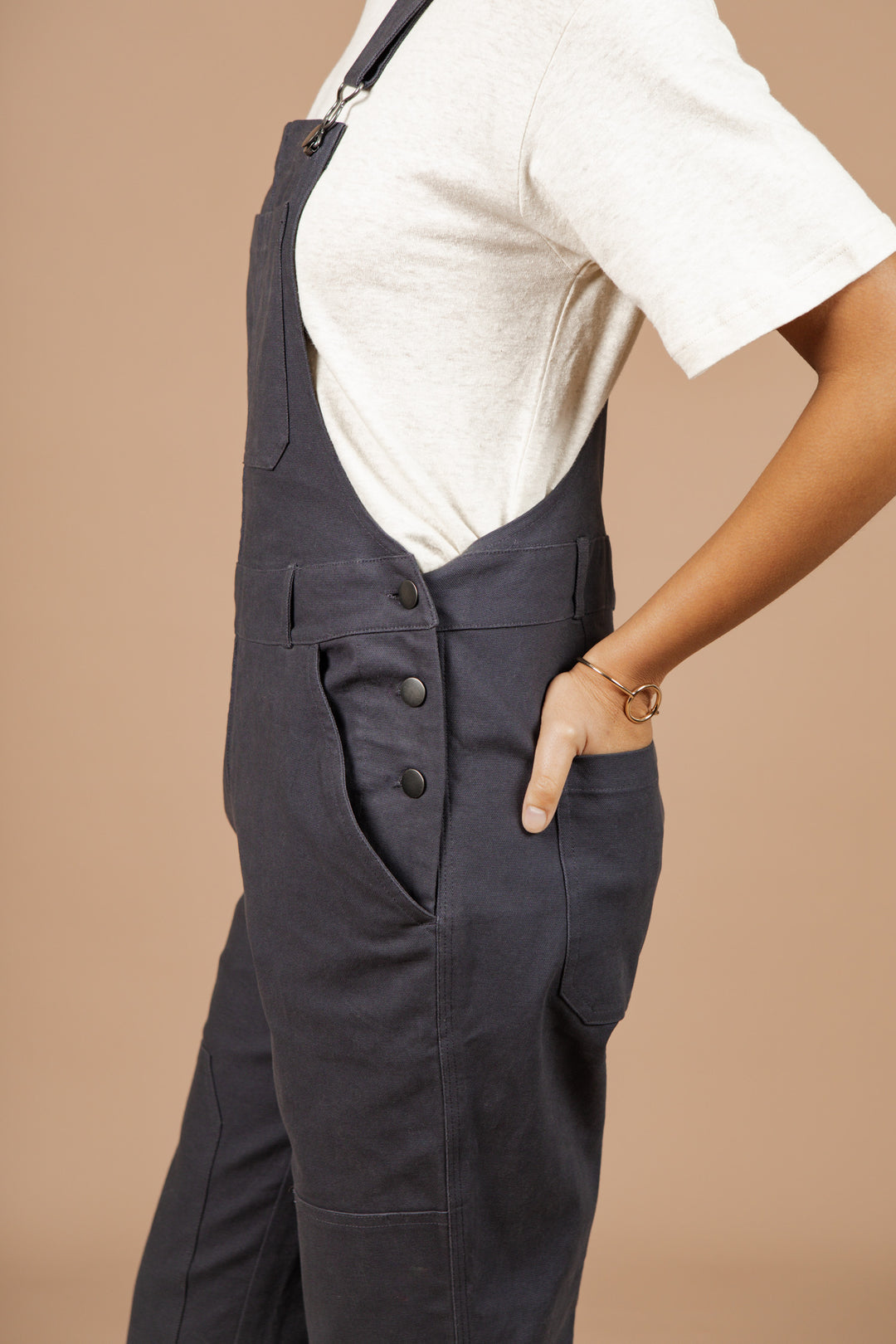 A person stands sideways with one hand in the pocket of Bridge & Burn's Tobin Utility Overall in Dark Charcoal Canvas, paired with a beige short-sleeve shirt and gold bracelet. The straight-leg overalls stand out against a plain light brown background.