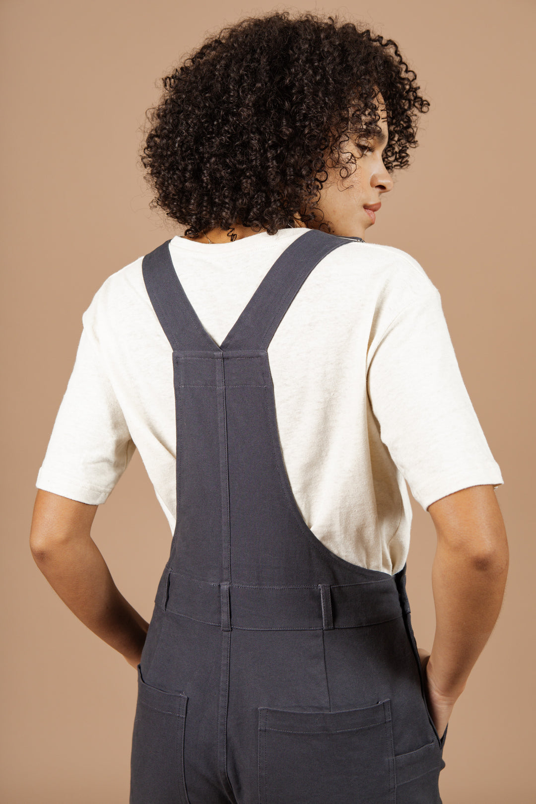 A person with curly hair, shown from the back against a beige background, wears a cream short-sleeve shirt and the Bridge & Burn Tobin Utility Overall in Dark Charcoal Canvas.