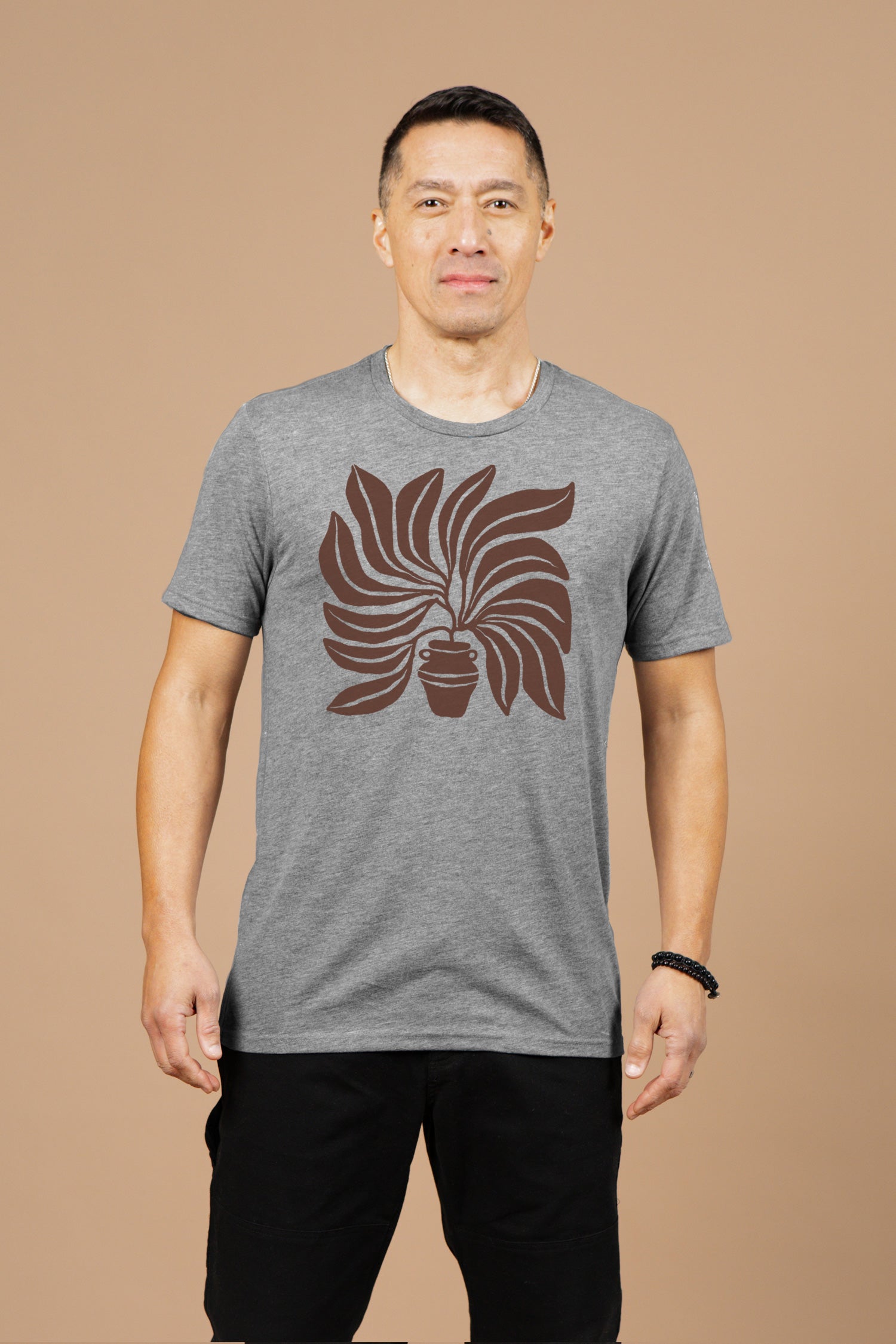 A man stands against a beige background, wearing Bridge & Burn’s Men's Grown Tee in grey featuring a large brown potted plant graphic on the front, paired with black shorts.