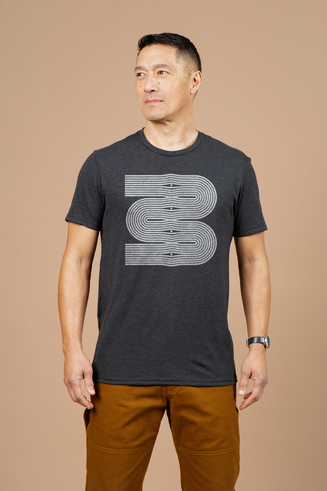 A man with short dark hair wears the Bridge & Burn Men's Side Winder Tee in black, featuring a large stylized "B" on the front. He stands against a plain beige background, paired with brown pants and a watch.
