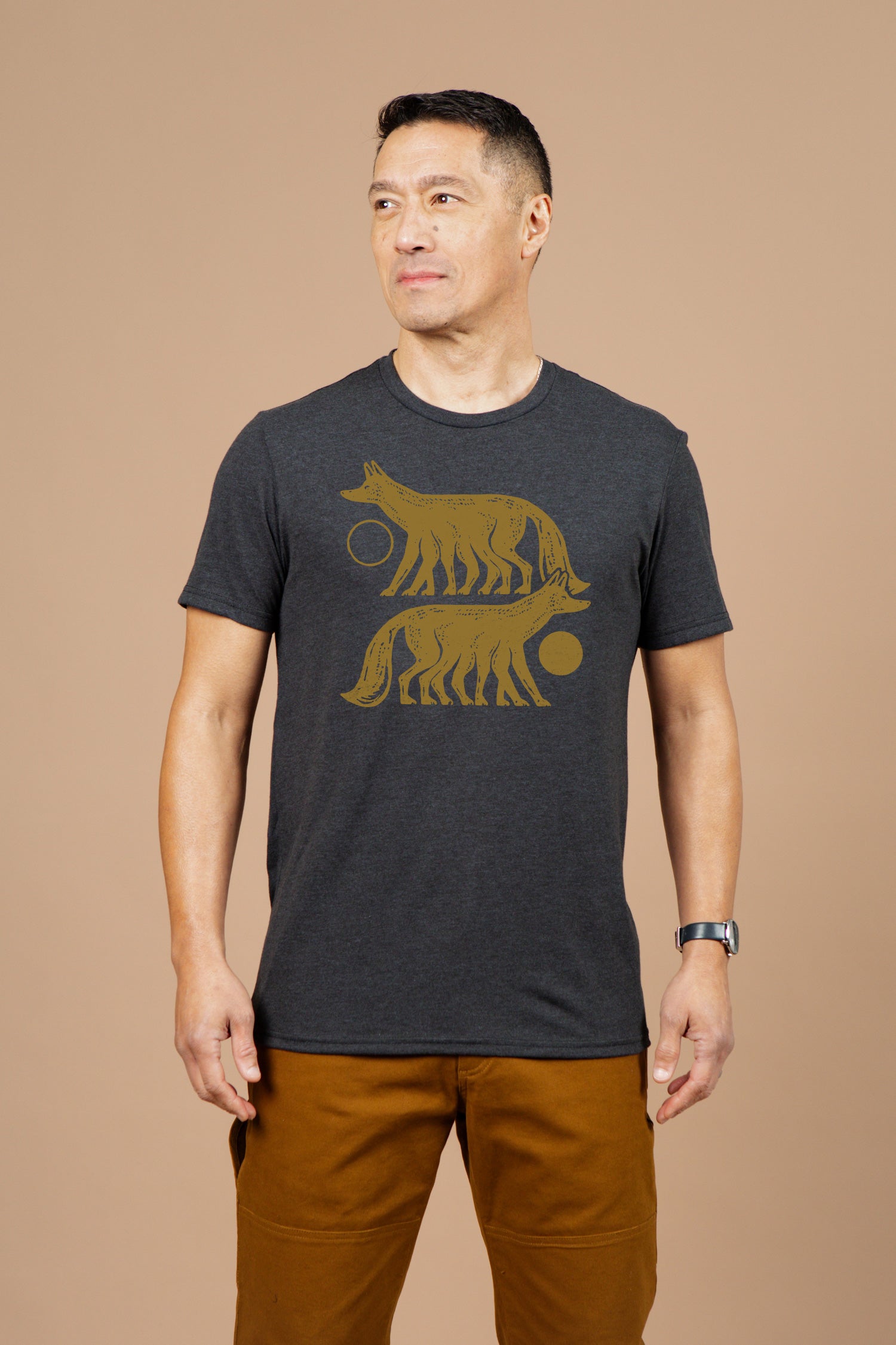 A man with short dark hair wears the Bridge & Burn Men's Night Watch Tee in black, featuring a gold graphic of two stylized wolves and circular shapes. He stands sideways against a beige background, paired with brown pants and a wristwatch.