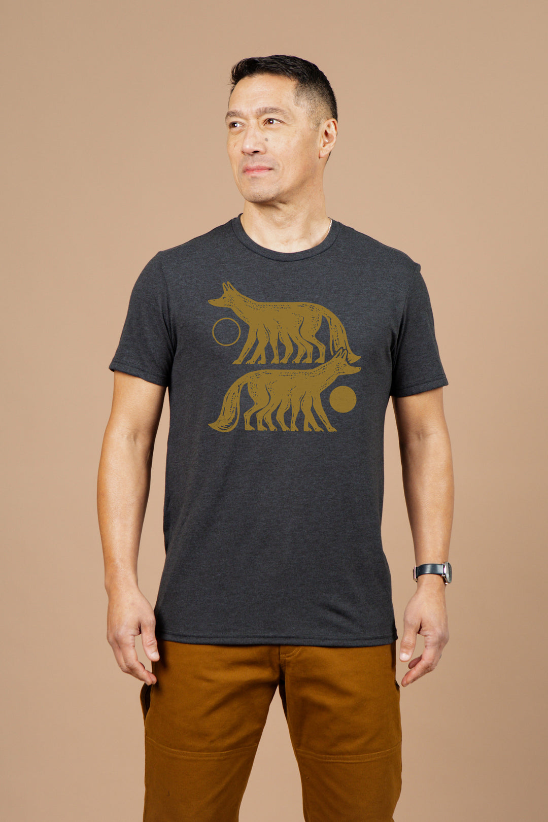 A man with short dark hair wears the Bridge & Burn Men's Night Watch Tee in black, featuring a gold graphic of two stylized wolves and circular shapes. He stands sideways against a beige background, paired with brown pants and a wristwatch.