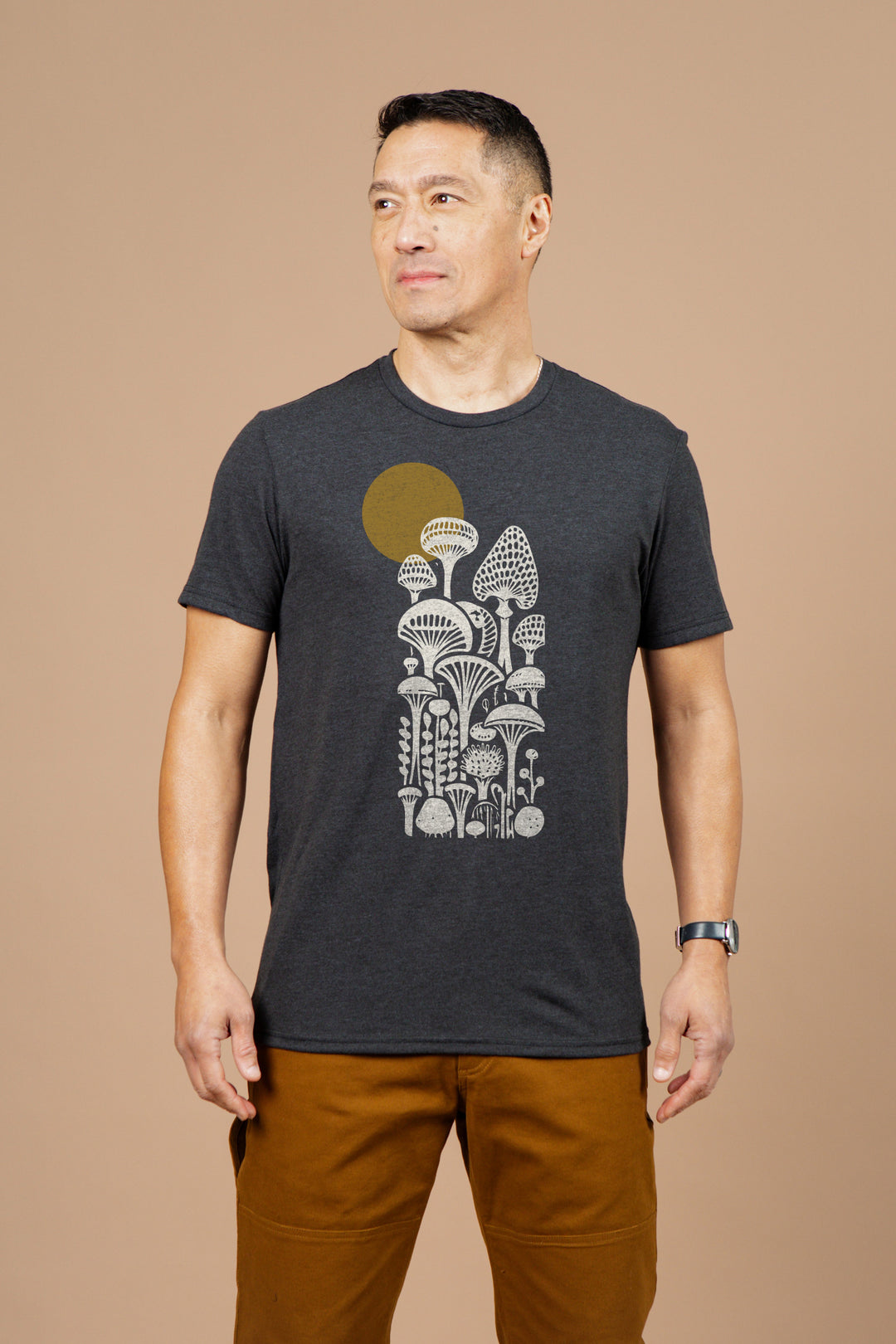 A man wears the Bridge & Burn Men's Mycology Tee in black, featuring white illustrated mushrooms and a yellow circle, paired with brown pants, as he stands against a plain beige background.
