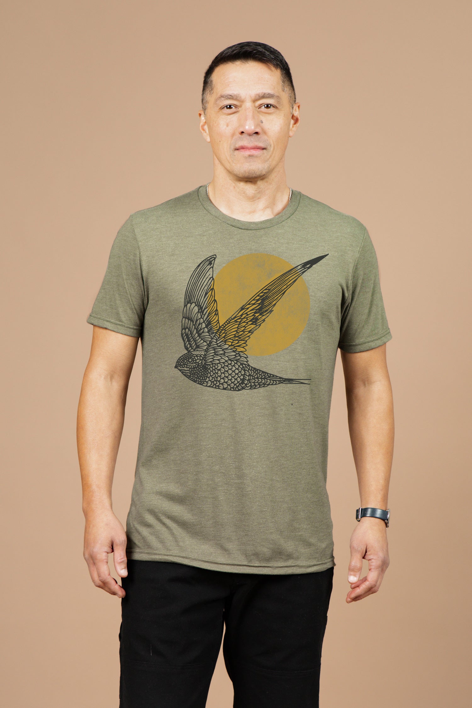 A man wears the Bridge & Burn Men's Flown Tee in olive, featuring a large bird and yellow sun graphic. He stands against a tan background, styled with short hair, black watch, and black pants.