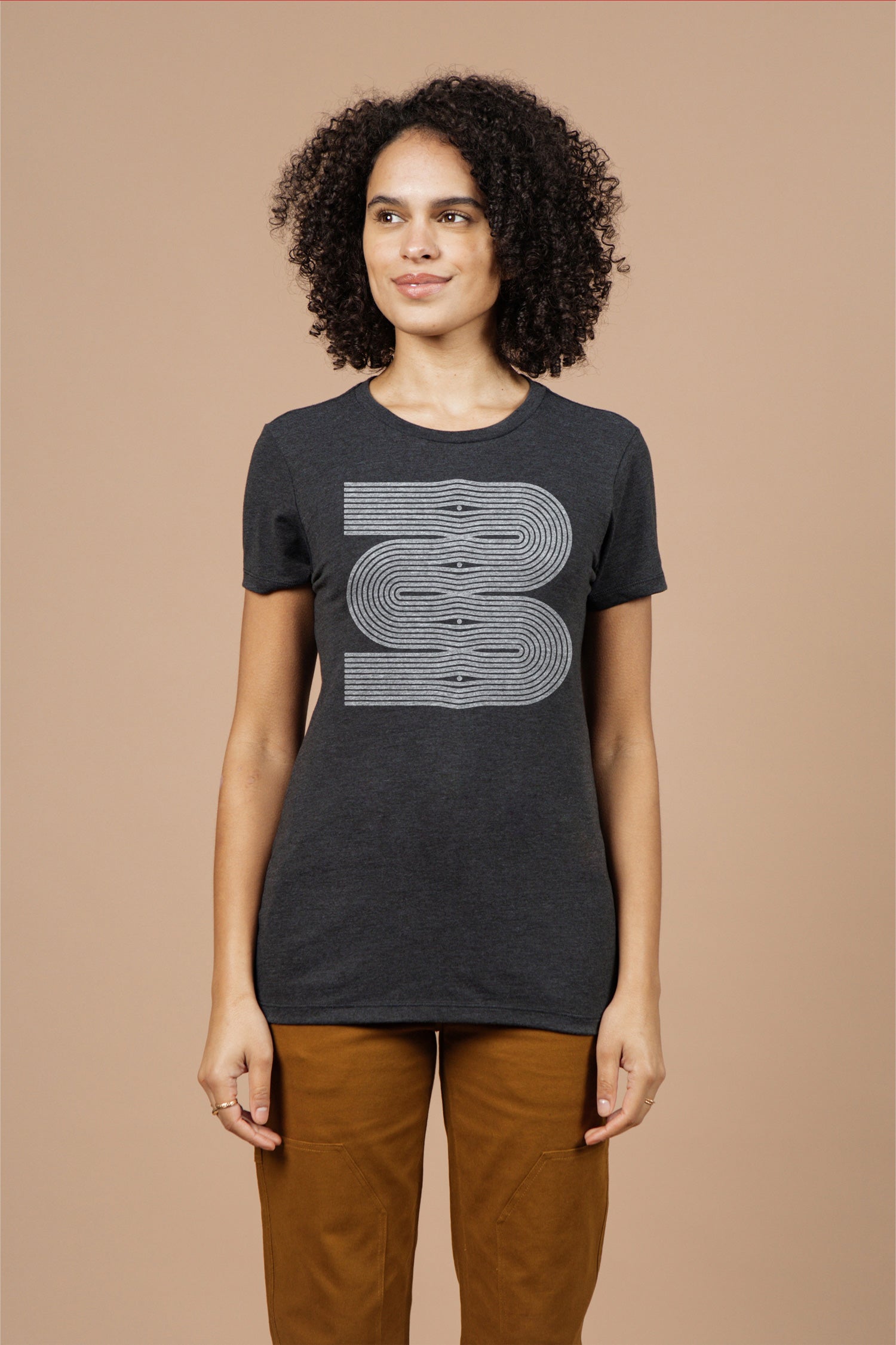 A person with curly hair wears the Bridge & Burn Women's Side Winder Tee in black, featuring a large, stylized number 3 in white lines. She pairs it with light brown pants and stands against a plain beige background.