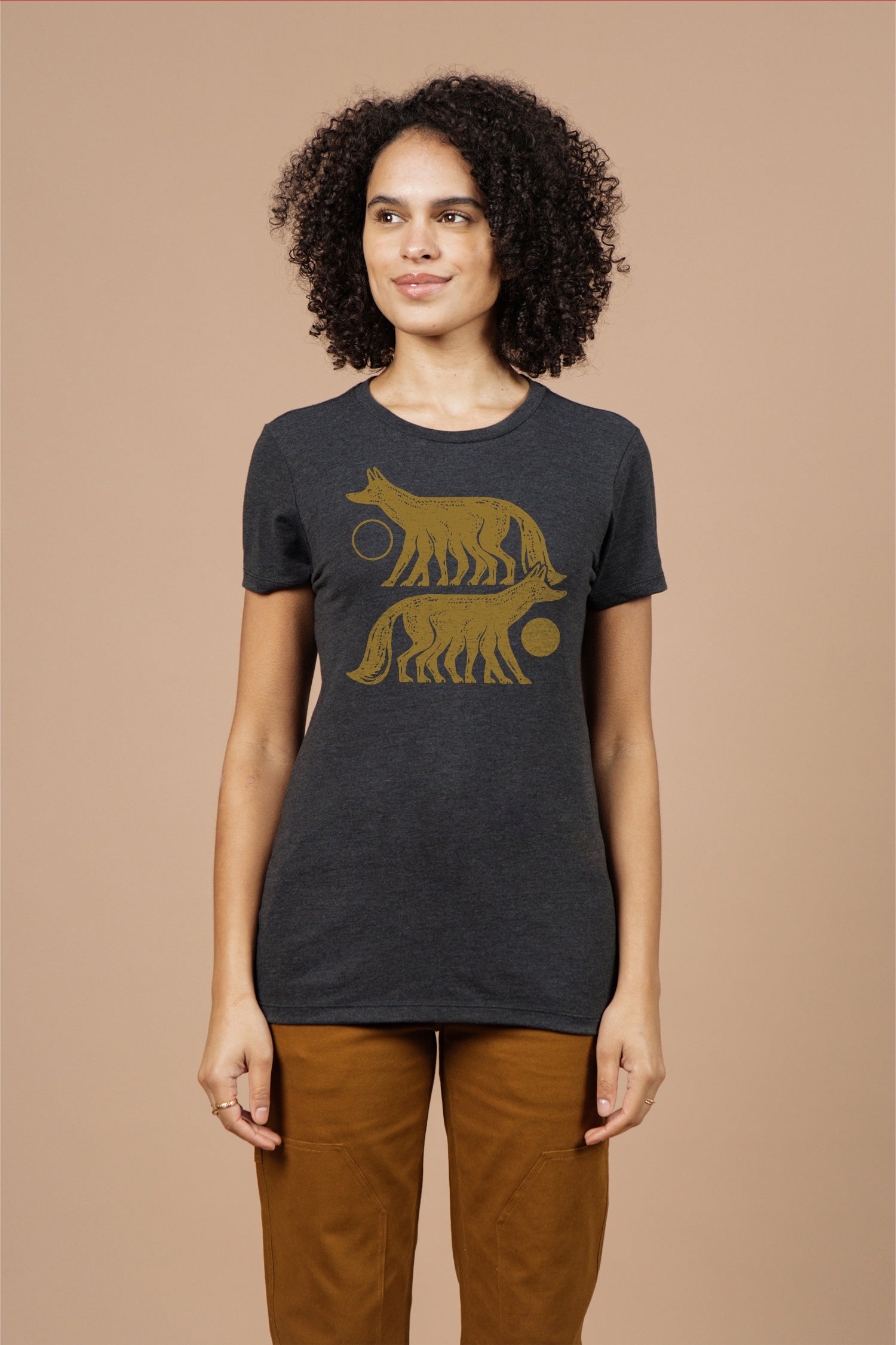 A woman with curly hair wears the Bridge & Burn Women's Night Watch Tee in black, featuring two gold wolf illustrations and circular designs. She stands against a beige background, looking slightly to the side with a soft smile.