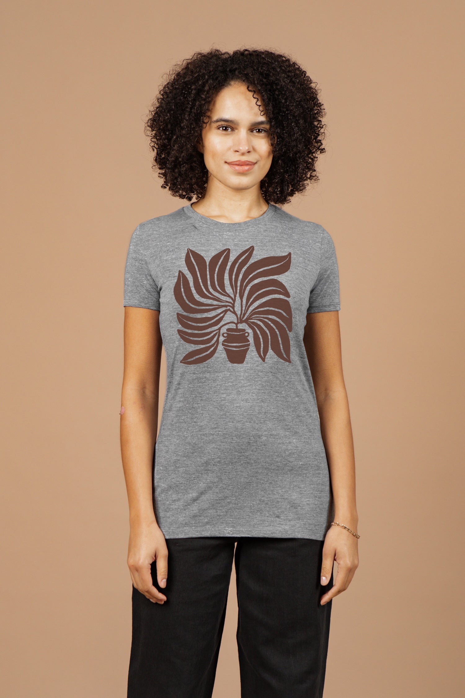 A woman with curly hair stands against a beige background, facing forward and smiling slightly in the Bridge & Burn Women's Grown Tee in grey, featuring organic cotton and a brown potted plant graphic, paired with black pants.
