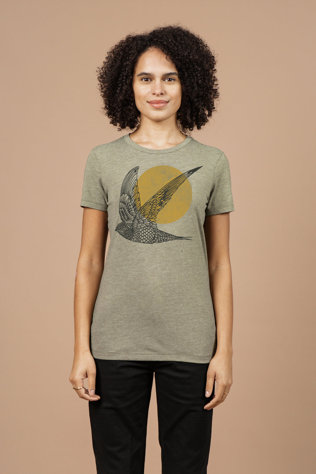 A person with curly hair wears Bridge & Burn's Women's Flown Tee in olive, an organic cotton shirt with a large bird graphic inside a yellow circle. The plain beige background highlights the design.