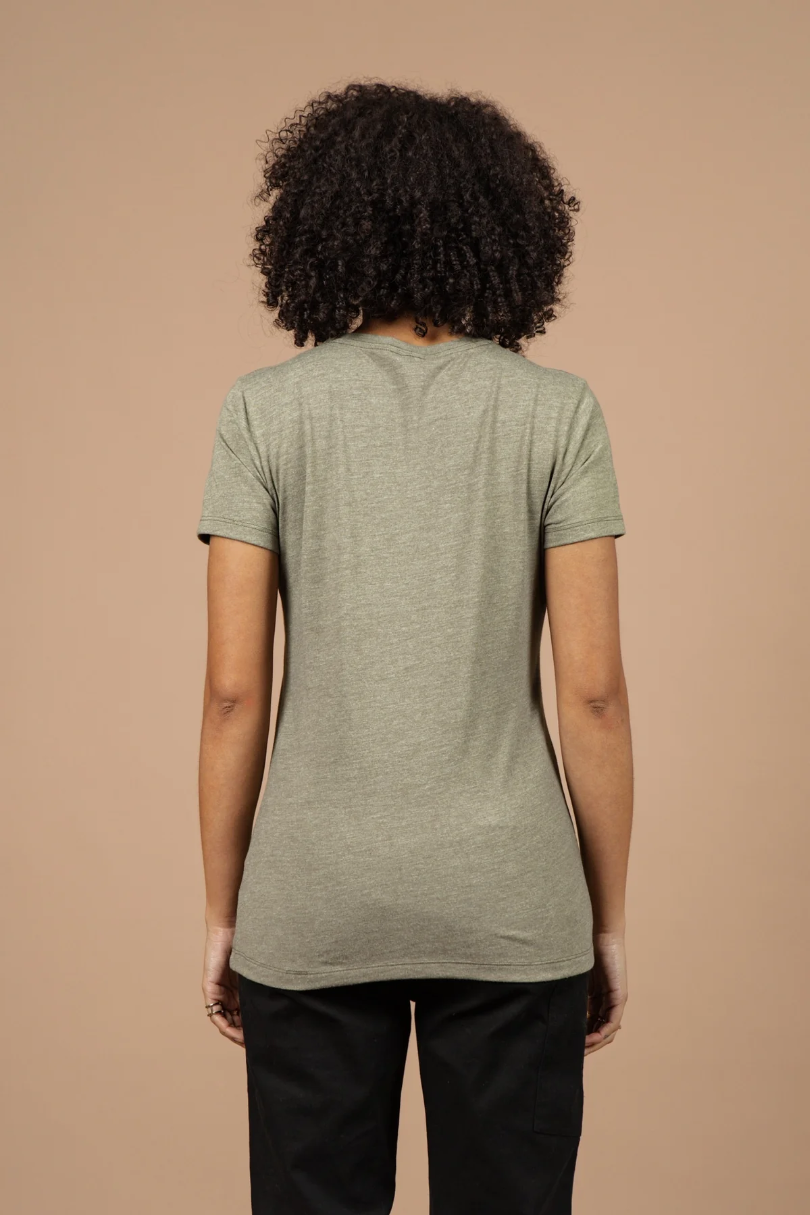 A woman with curly hair, wearing the Bridge & Burn Women's Flown Tee in olive and black pants, stands facing away from the camera against a plain beige background.