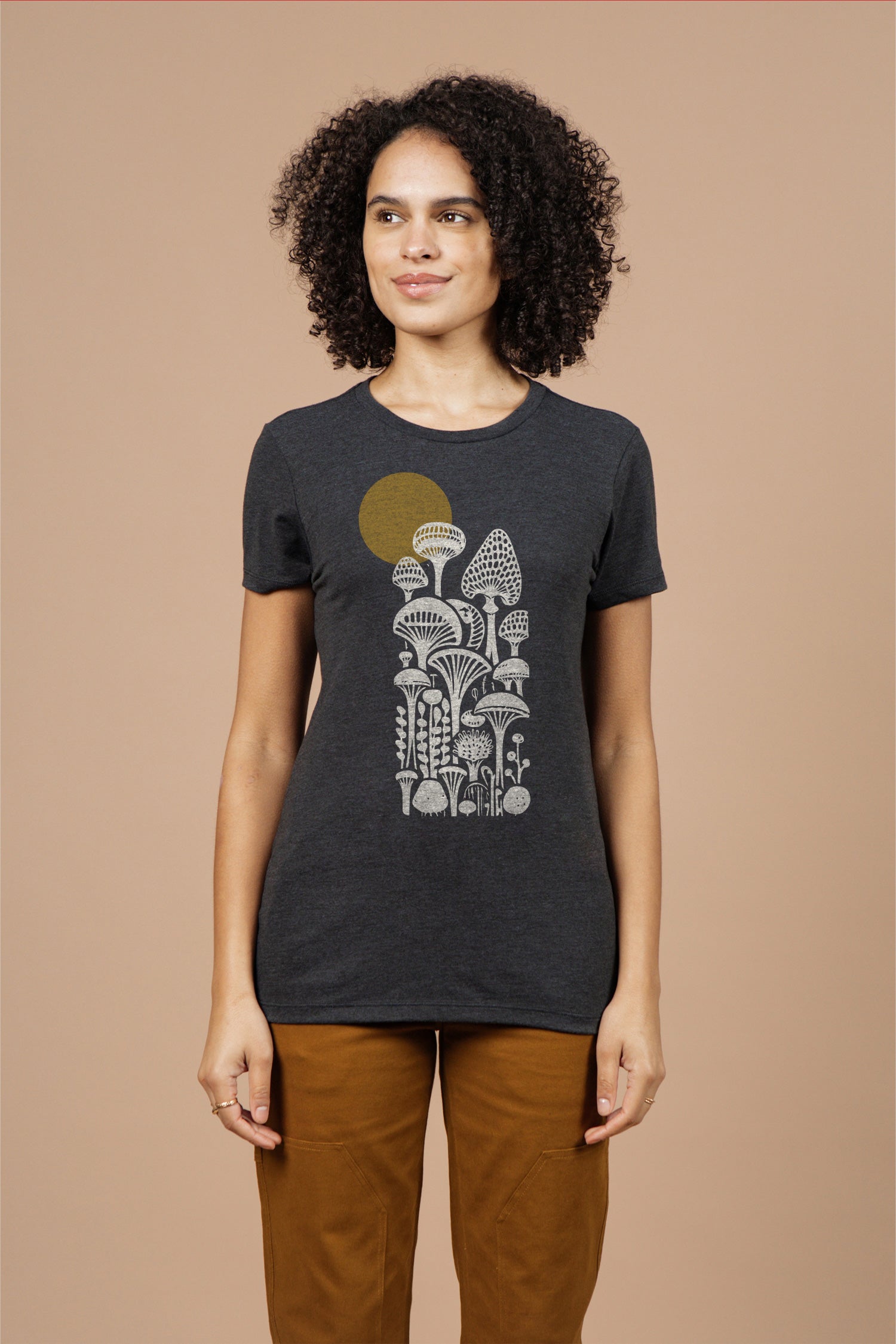 A woman wears the Bridge & Burn Women's Mycology Tee in black, featuring a graphic of mushrooms and a yellow sun, standing against a plain beige background.