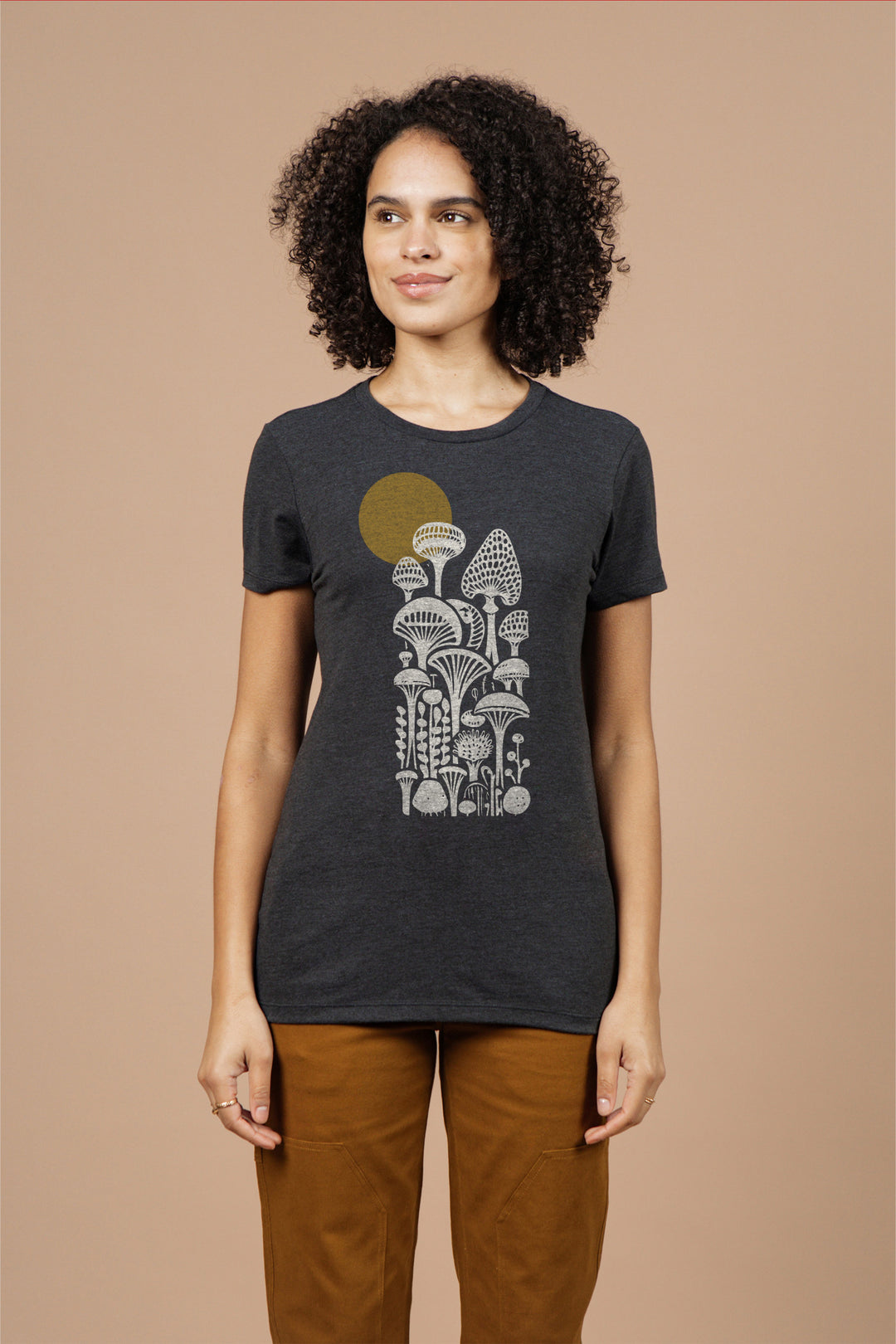 A woman wears the Bridge & Burn Women's Mycology Tee in black, featuring a graphic of mushrooms and a yellow sun, standing against a plain beige background.