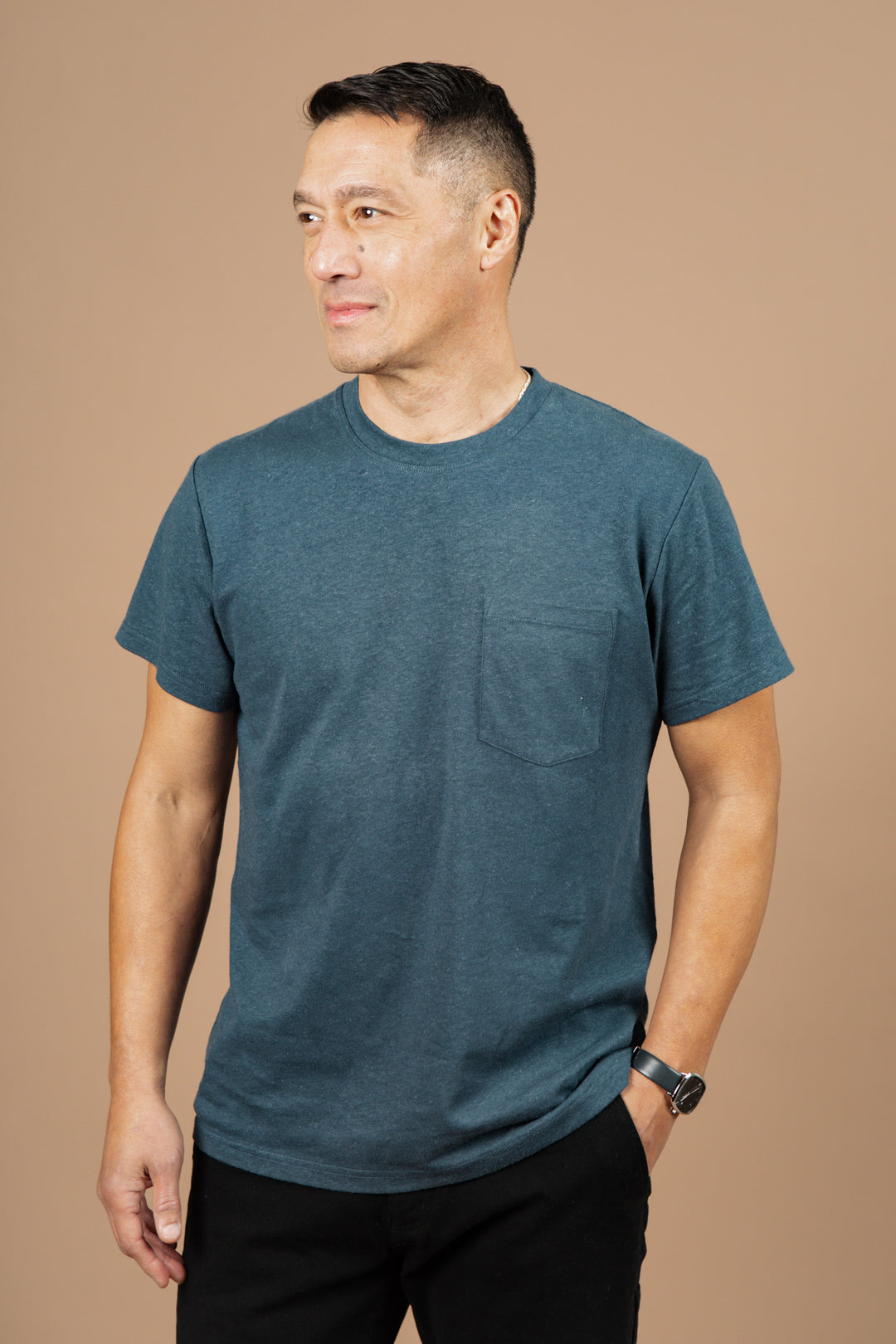 A man with short dark hair, wearing the Bridge & Burn Organic Hemp Pocket Tee in Storm Blue and black pants, stands against a plain beige background, looking slightly to his left.