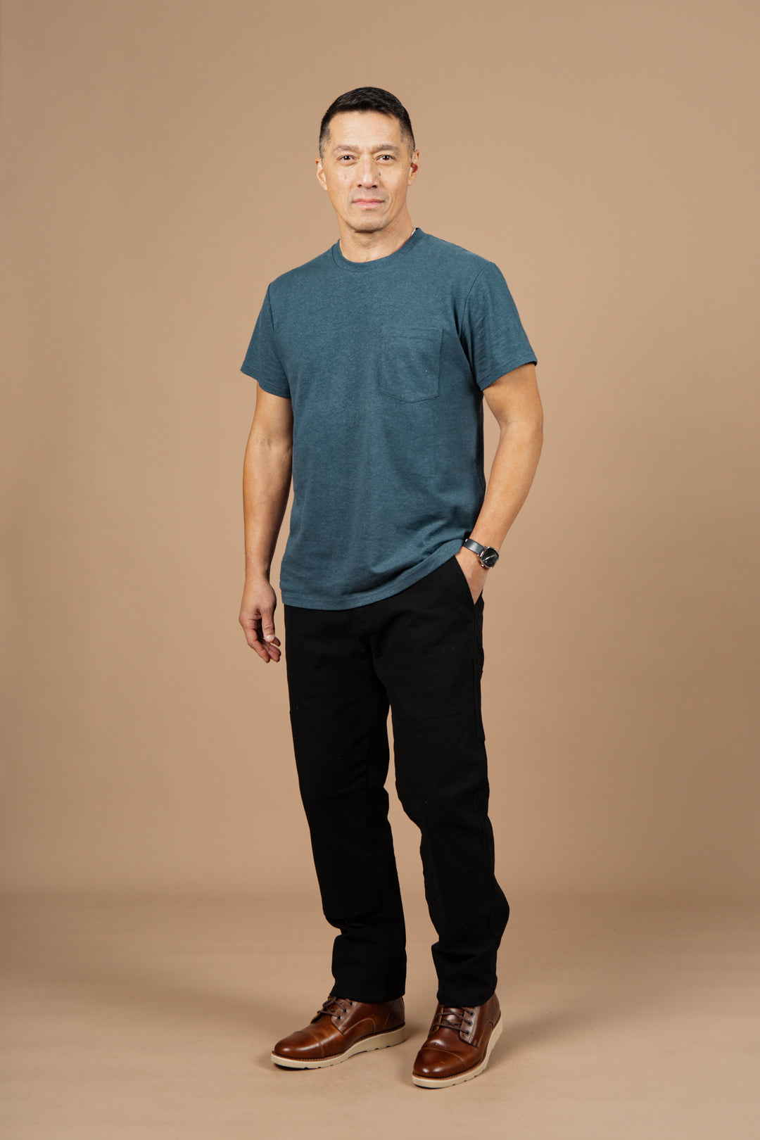 A man stands against a plain beige background, wearing the Bridge & Burn Organic Hemp Pocket Tee in Storm Blue, black pants, brown leather shoes, a watch, and has one hand in his pocket with a neutral expression.