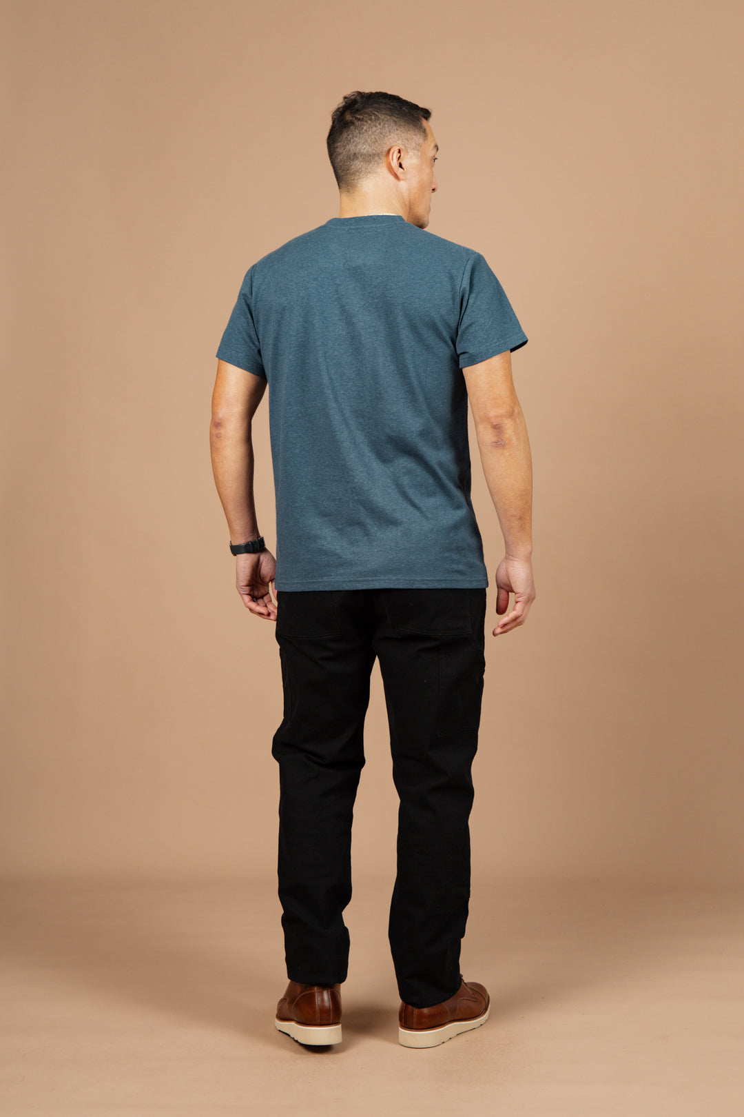 A man stands facing away from the camera, wearing the Bridge & Burn Organic Hemp Pocket Tee in Storm Blue, black pants, brown shoes, and a watch against a plain beige background.