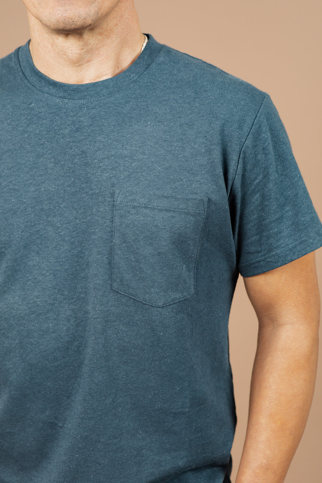 A person wears the Bridge & Burn Organic Hemp Pocket Tee in Storm Blue, featuring a chest pocket. Only their upper torso and part of their arms are visible against a plain beige background.