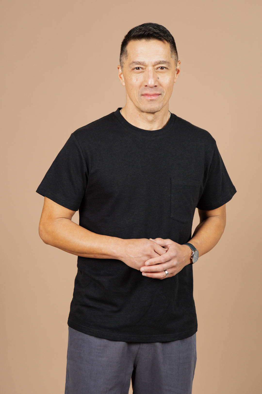 A man with short dark hair stands against a beige background, wearing the Bridge & Burn Organic Hemp Pocket Tee in Washed Black and gray pants. He has a neutral expression and his hands are clasped together in front of him.