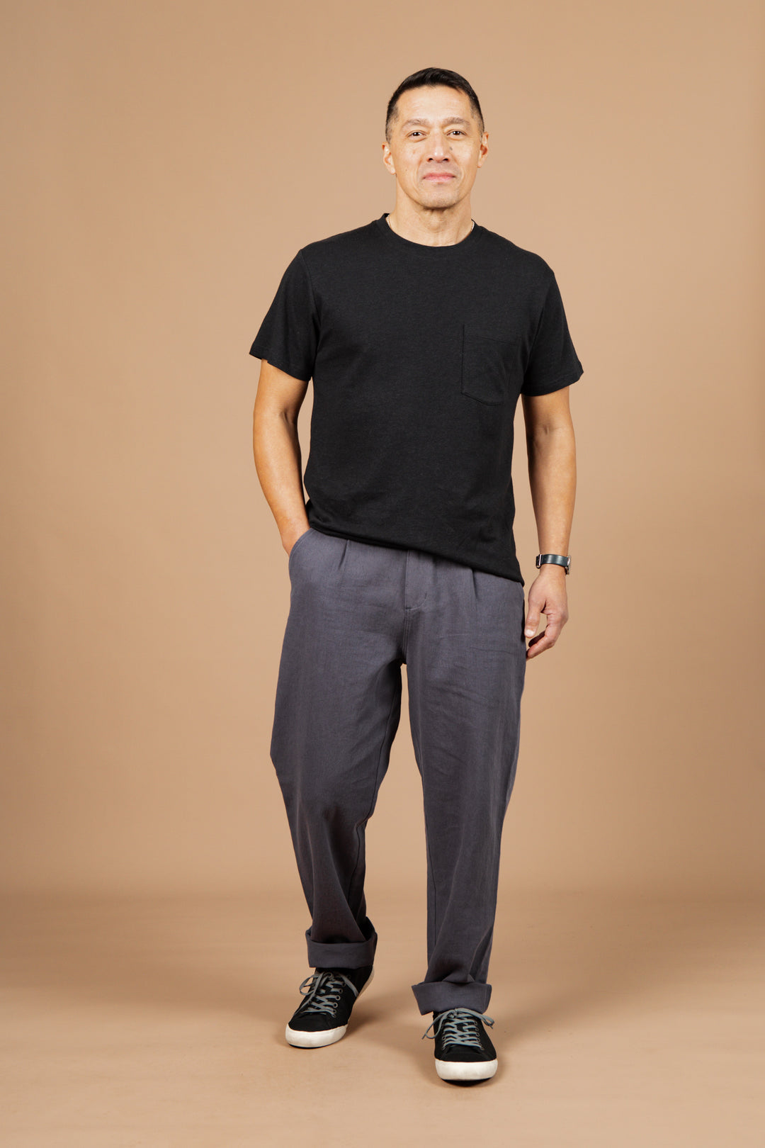 A man stands against a beige background wearing the Bridge & Burn Organic Hemp Pocket Tee in Washed Black, with gray pants, black sneakers, and a wristwatch. His hands are in his pockets as he faces the camera with a relaxed expression.