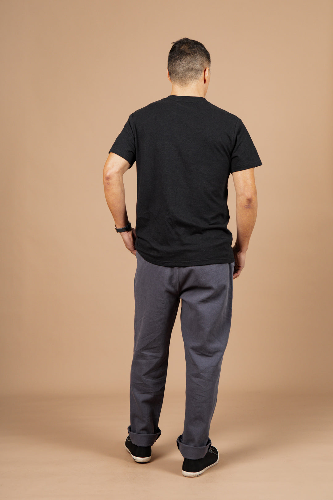 A man stands with his back to the camera, wearing the Bridge & Burn Organic Hemp Pocket Tee in Washed Black, paired with gray pants and black shoes, against a plain beige background.