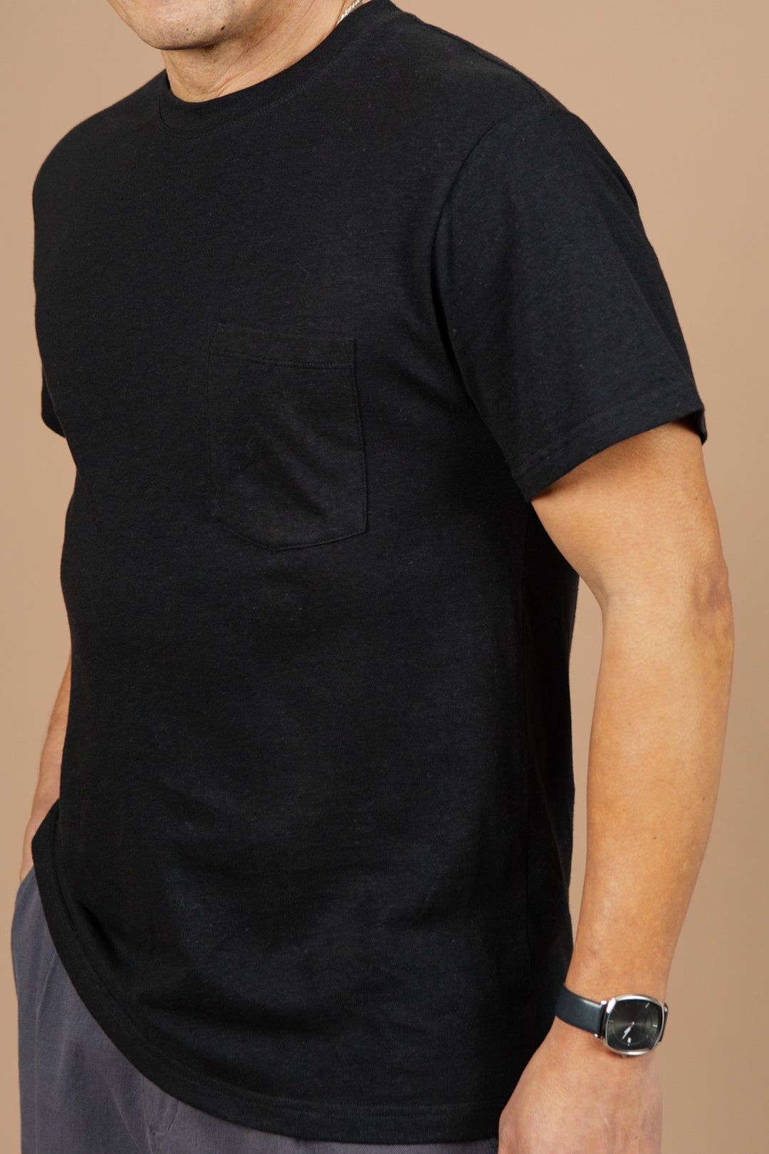 A person in a Bridge & Burn Organic Hemp Pocket Tee in Washed Black, gray pants, and a black wristwatch stands against a beige background with one hand in their pocket; their face is partially obscured.