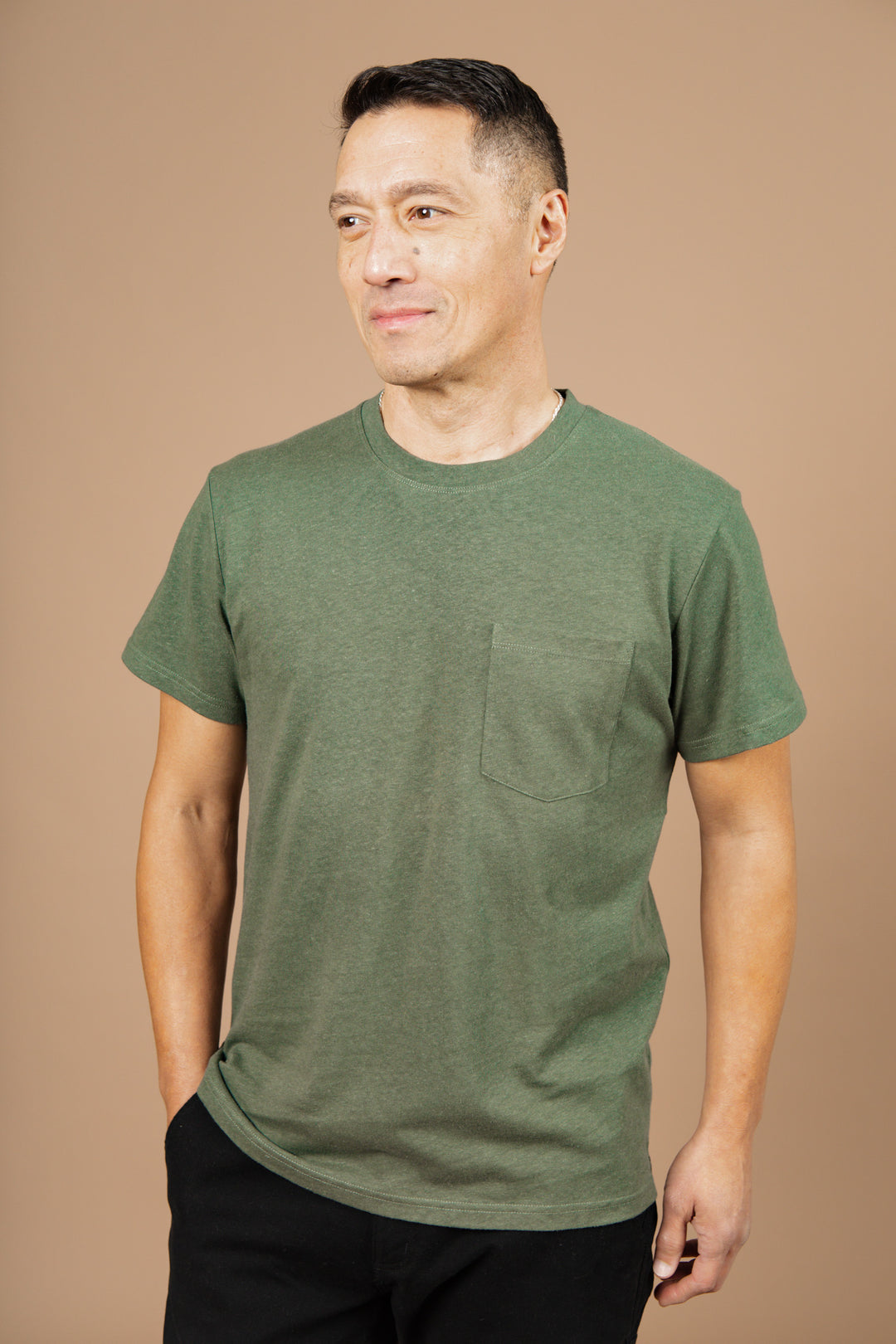 A man with short dark hair wears the Bridge & Burn Organic Hemp Pocket Tee in Cypress and black pants, standing against a plain beige background and looking slightly to the side with a neutral expression.