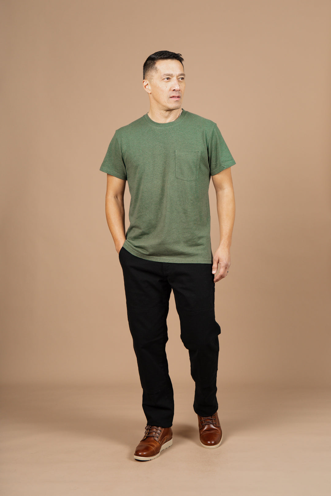 A man with short dark hair stands against a beige background, wearing Bridge & Burn's Organic Hemp Pocket Tee in Cypress, black pants, and brown shoes. He has one hand in his pocket and gazes slightly to the side.