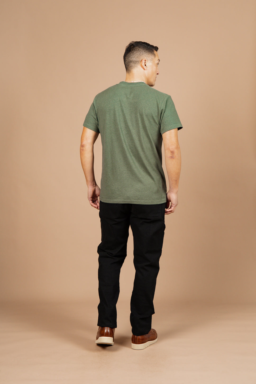 A man stands with his back to the camera against a beige background, wearing Bridge & Burn's Organic Hemp Pocket Tee in Cypress, along with black pants and brown shoes. His arms hang naturally at his sides.