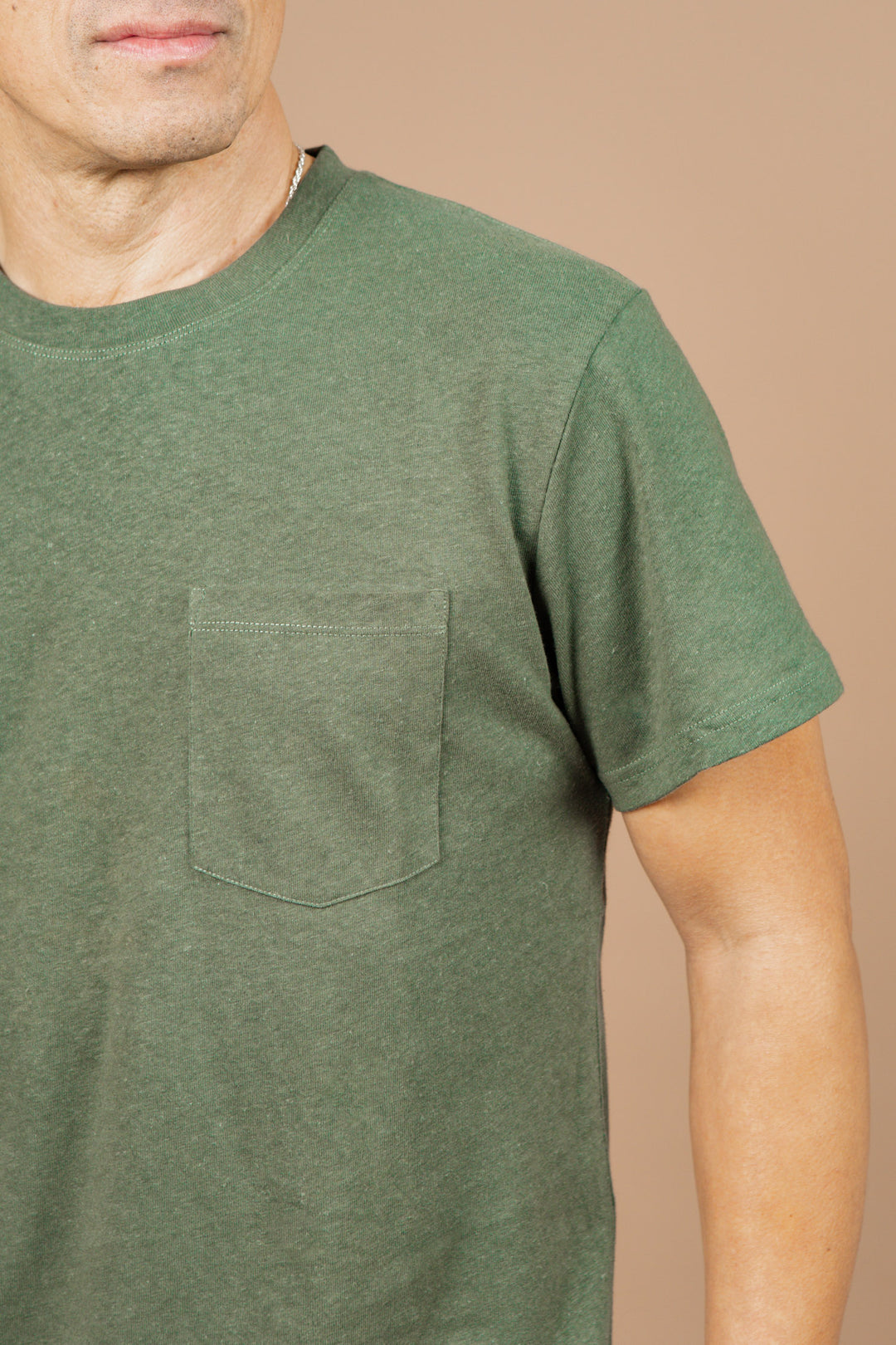 A man wears the Bridge & Burn Organic Hemp Pocket Tee in Cypress, featuring a chest pocket, standing against a beige background. Only his upper body and part of his face are visible.