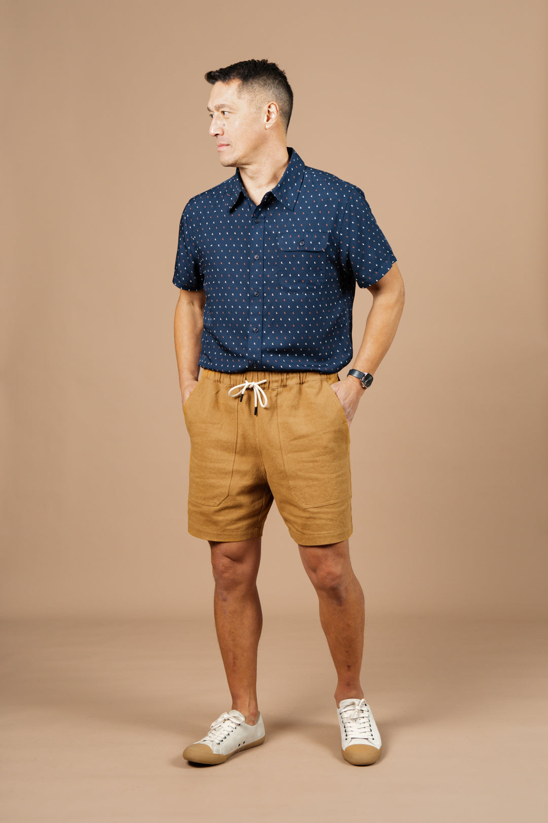 A man stands against a beige background wearing the Bridge & Burn Noah Short in Ochre with a relaxed fit, paired with a dark blue short-sleeved shirt with white dots, white sneakers, and a wristwatch. He has his hands in his pockets and looks to the side.