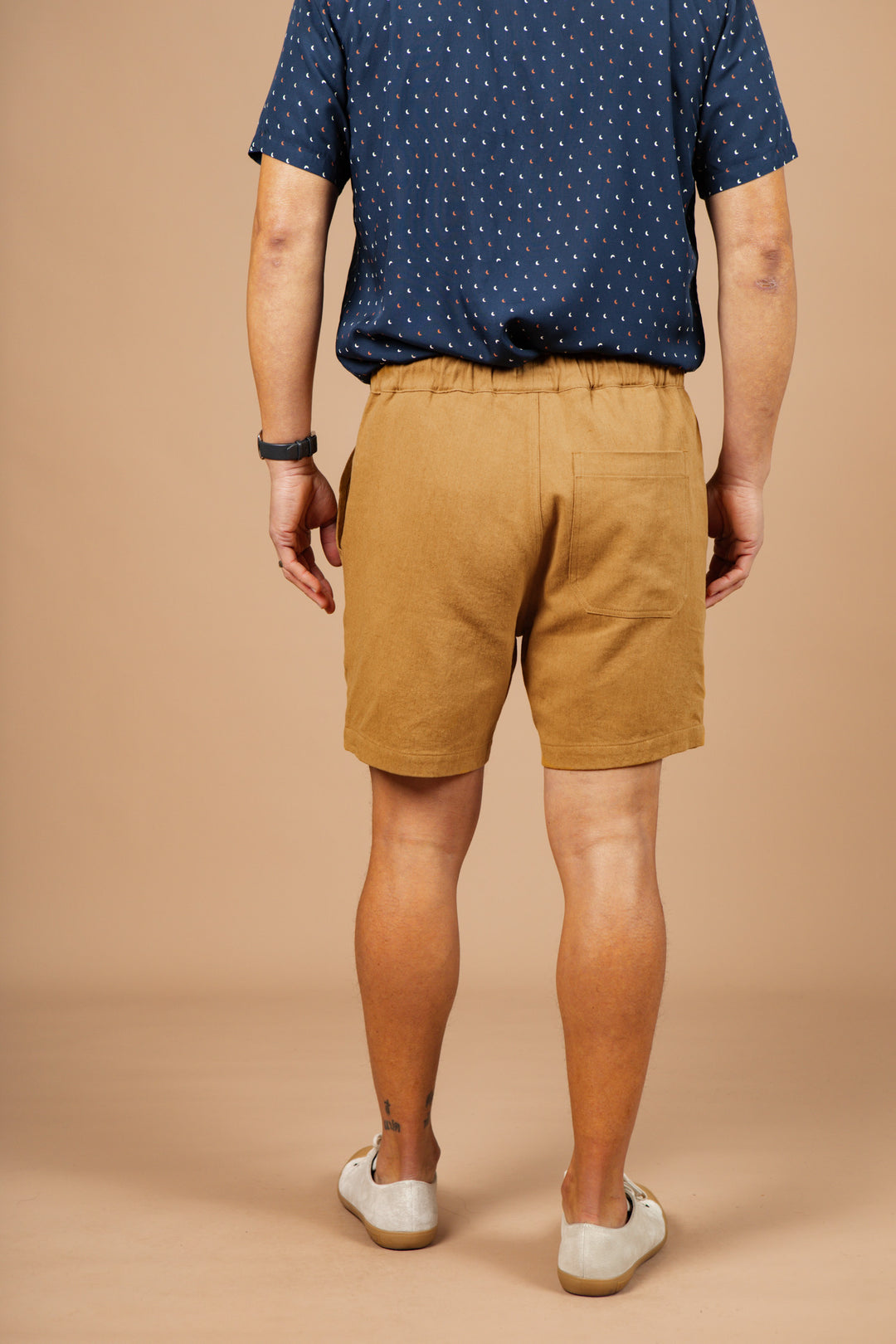 A person stands facing away, wearing Bridge & Burn's Noah Short in Ochre, a navy blue short-sleeve shirt with white dots, a black watch, and white slip-on shoes against a plain beige background.