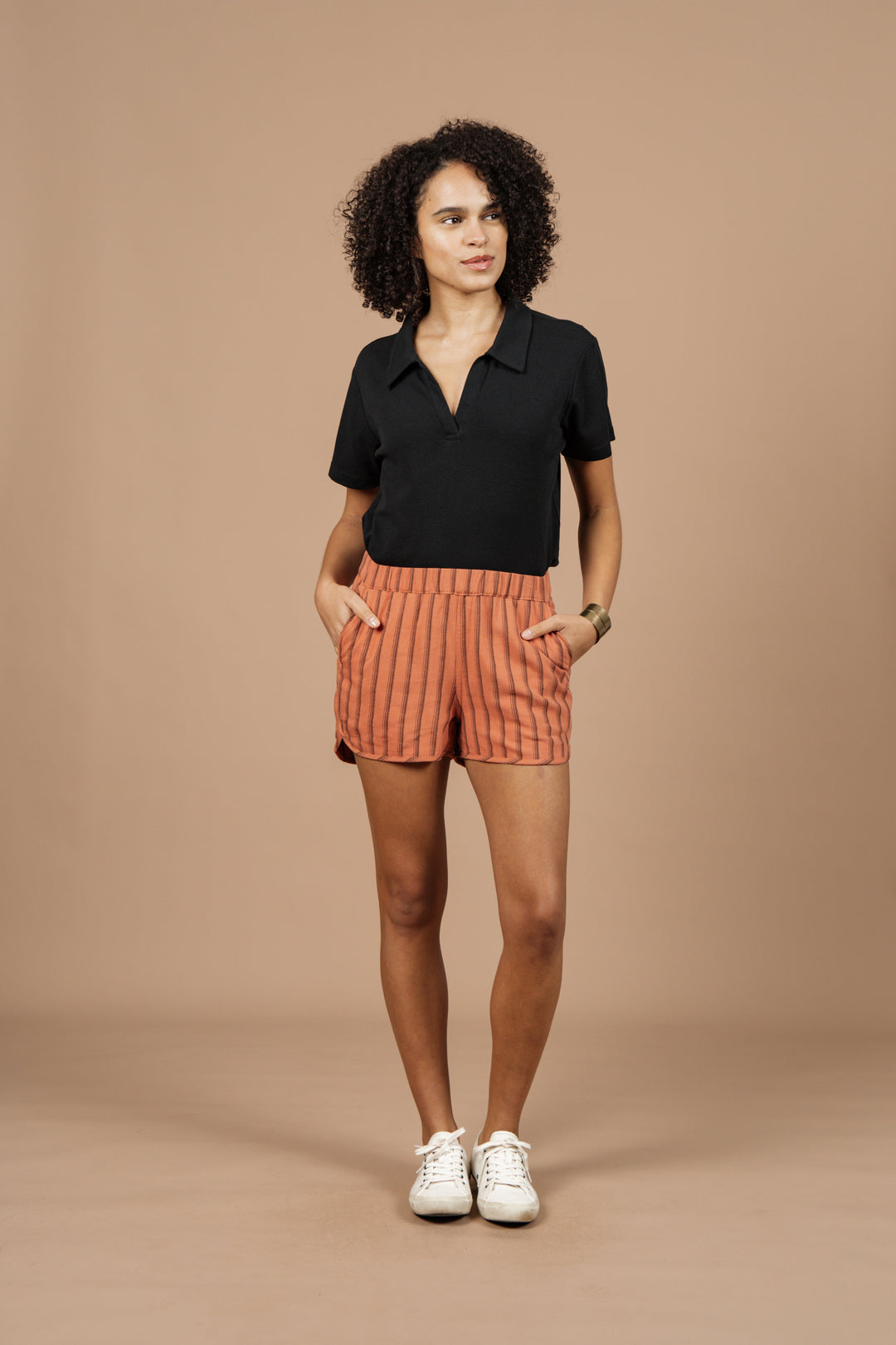 A woman stands against a tan background wearing Bridge & Burn's Luca Track Short in Rust Stripe, paired with a black short-sleeve polo, white sneakers, and a bracelet, with her hands in her pockets.