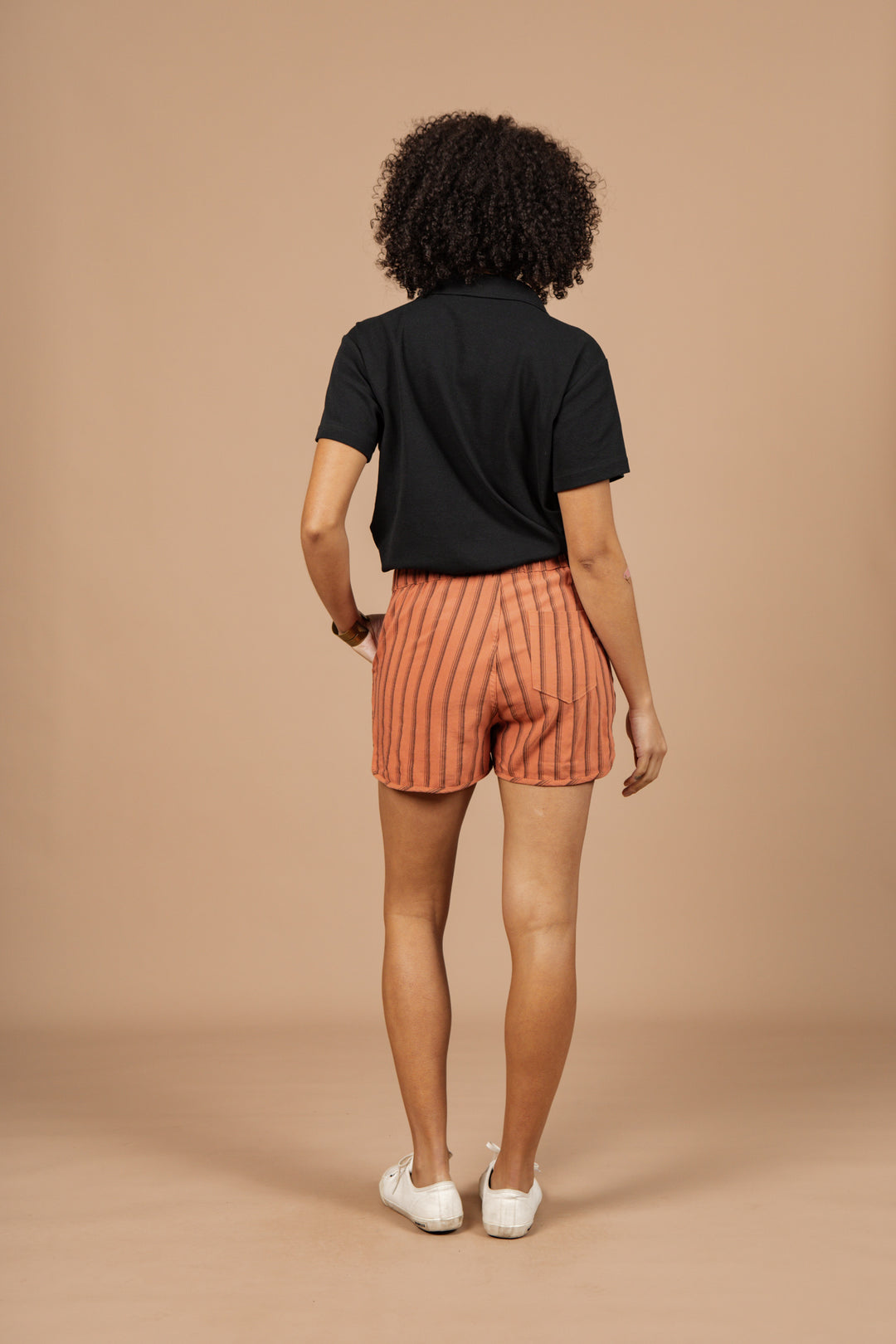 A person with curly hair, wearing a black shirt, Bridge & Burn Luca Track Short in Rust Stripe, and white shoes stands facing away from the camera against a plain beige background.