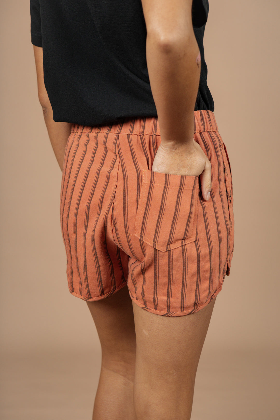 A person stands with one hand in the pocket of Bridge & Burn’s Luca Track Short / Rust Stripe, featuring vertical black stripes, paired with a black top against a plain beige background.