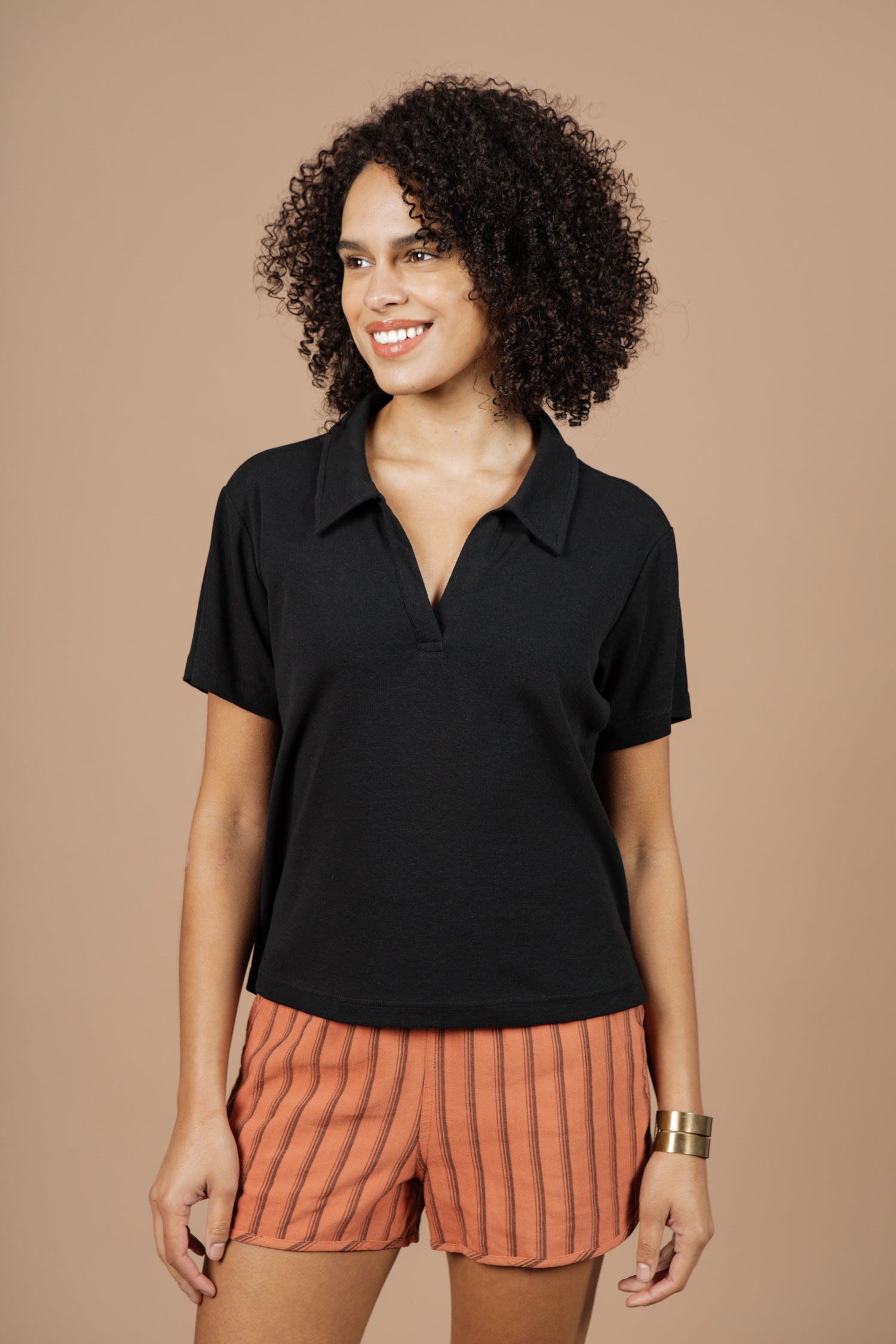 A person with curly hair stands smiling against a plain beige background, wearing the Bridge & Burn Avery Boxy Top in black jersey and orange striped shorts.