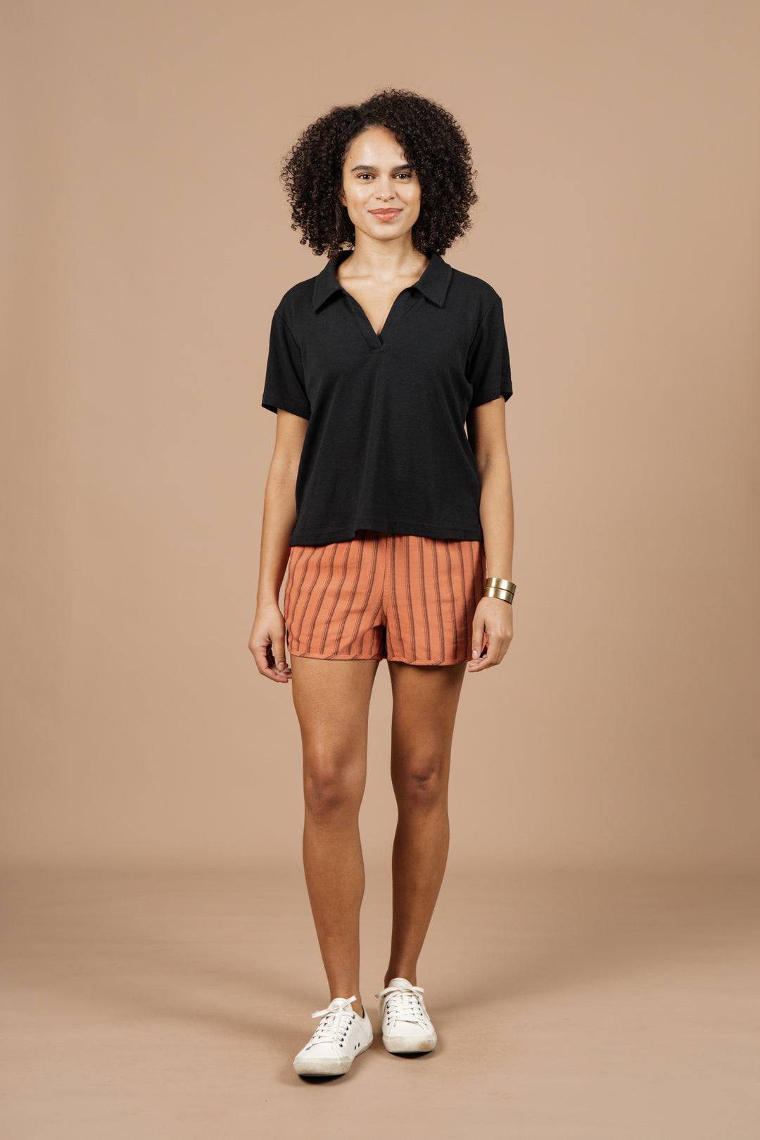 A person stands against a plain beige background, smiling with hands relaxed at their sides, wearing the Bridge & Burn Avery Boxy Top in black jersey, orange striped shorts, white sneakers, and a gold bracelet.