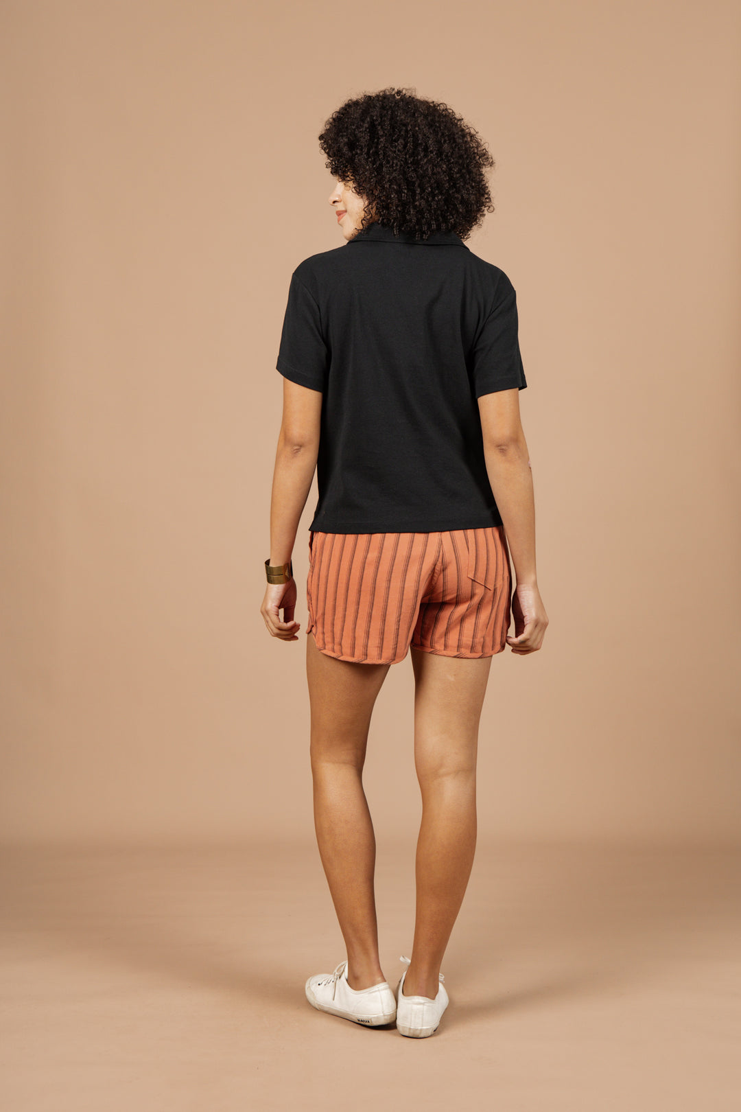 A person with curly hair faces away, wearing the Bridge & Burn Avery Boxy Top in black jersey, orange striped shorts, white shoes, and a bracelet. The background is solid beige.