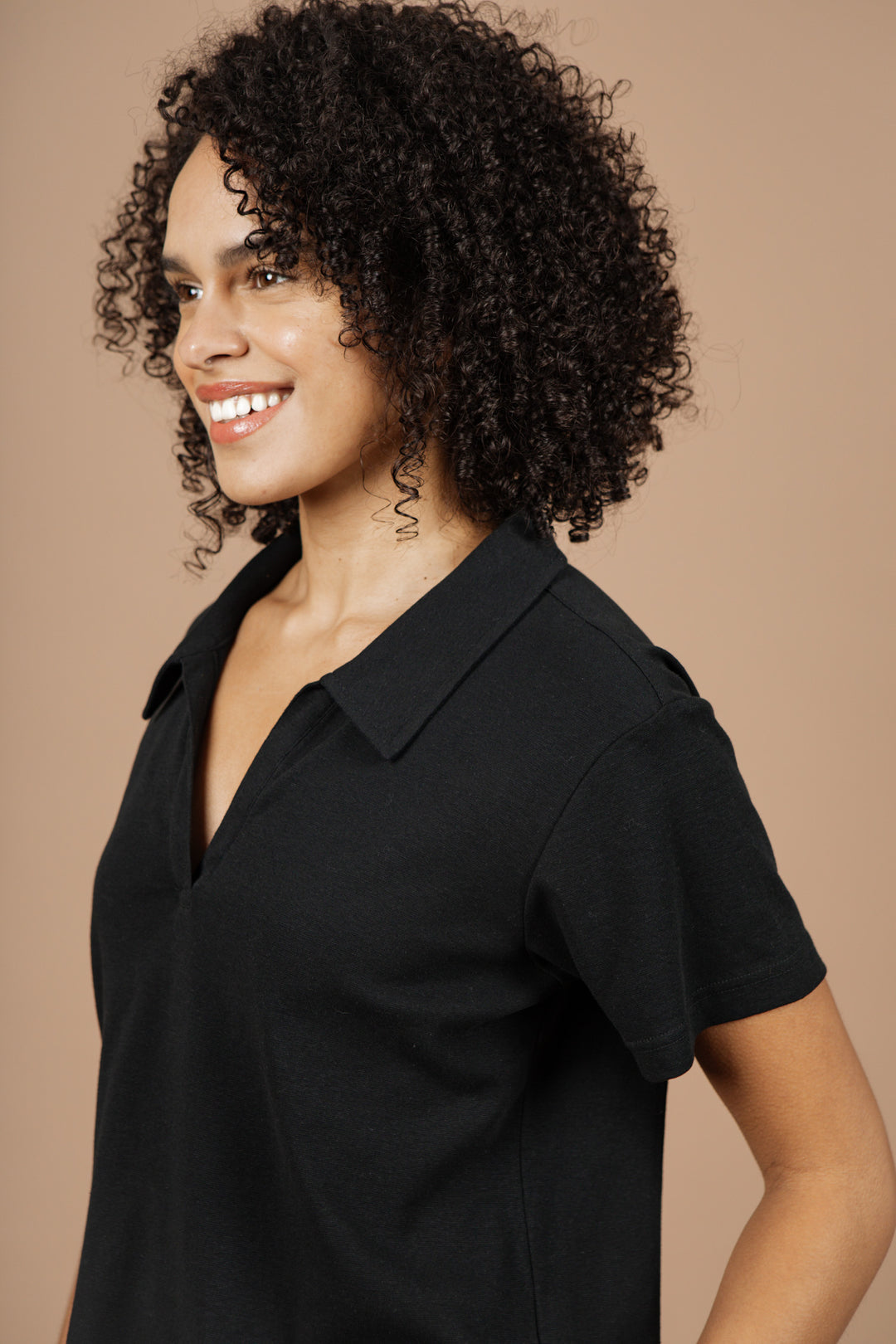 A woman with curly hair and a bright smile wears the Bridge & Burn Avery Boxy Top in black jersey, posing against a plain beige background.