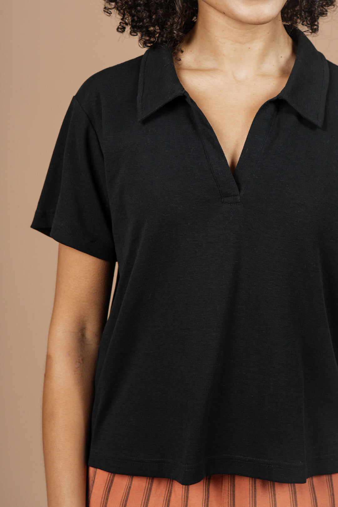 A person wearing the Bridge & Burn Avery Boxy Top in black jersey stands against a plain beige background. Their face is partially out of the frame, and they pair the top with orange striped pants.