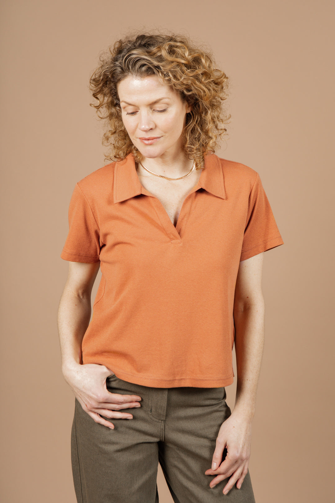A woman with curly blonde hair wears the Bridge & Burn Avery Boxy Top in clay red jersey and olive green pants, standing against a plain beige background and looking down with a slight smile.