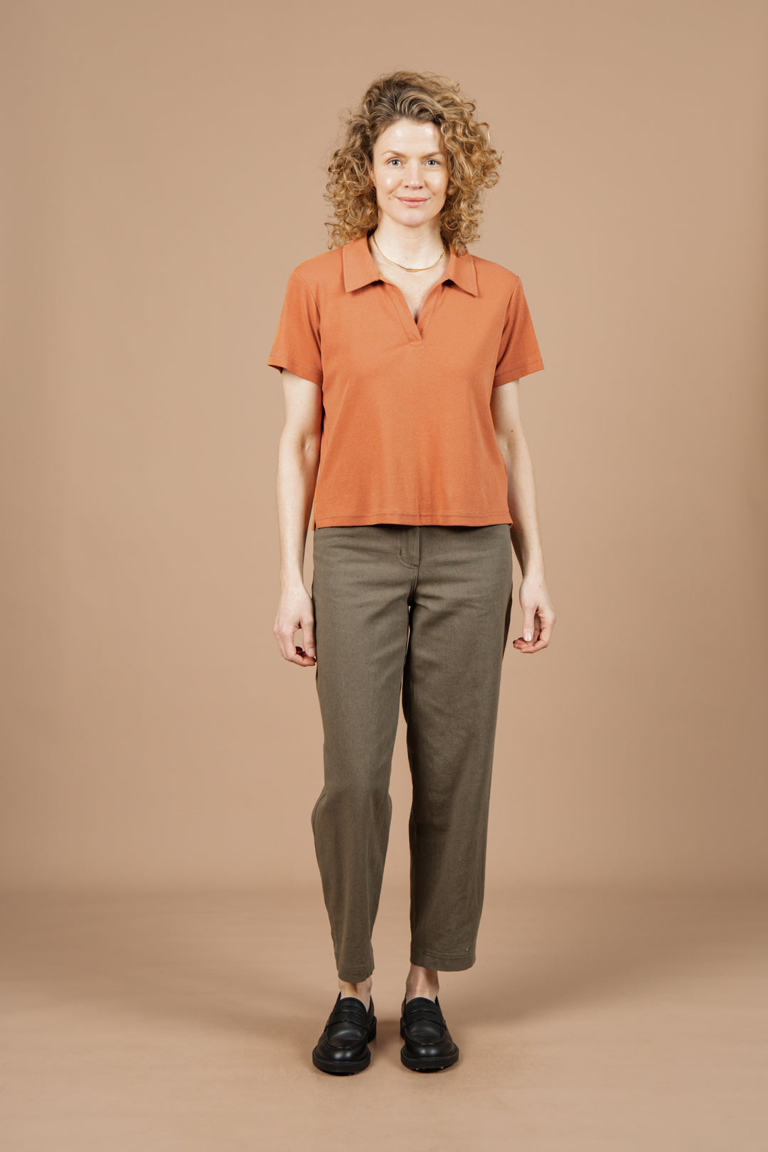 A person with curly hair wears the Bridge & Burn Avery Boxy Top in Clay Red Jersey, along with olive green pants and black shoes, standing facing the camera against a plain beige background.