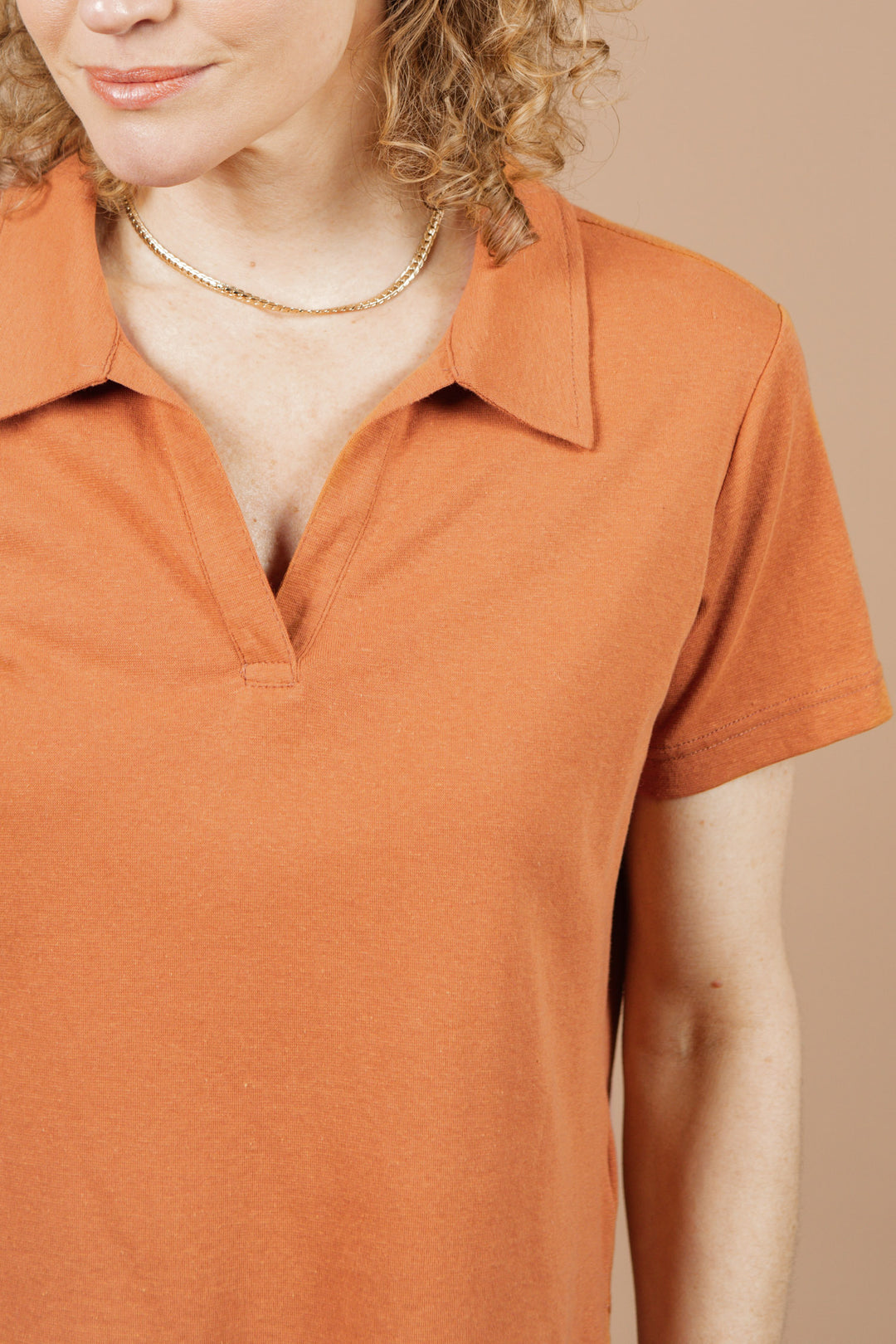 A woman wears the Bridge & Burn Avery Boxy Top in Clay Red Jersey, styled with a gold chain necklace. The photo shows her from shoulders to nose against a beige background.