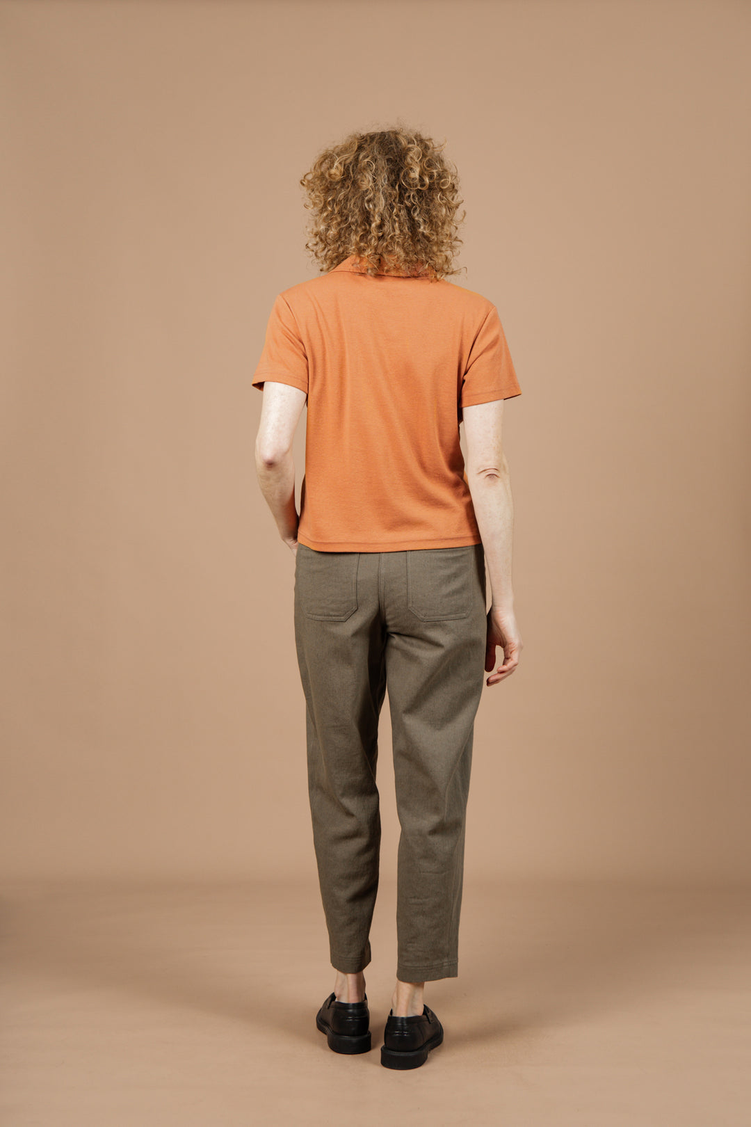 A person with curly hair, wearing the Bridge & Burn Avery Boxy Top in Clay Red Jersey, olive green pants, and black shoes stands facing away from the camera against a plain beige background.