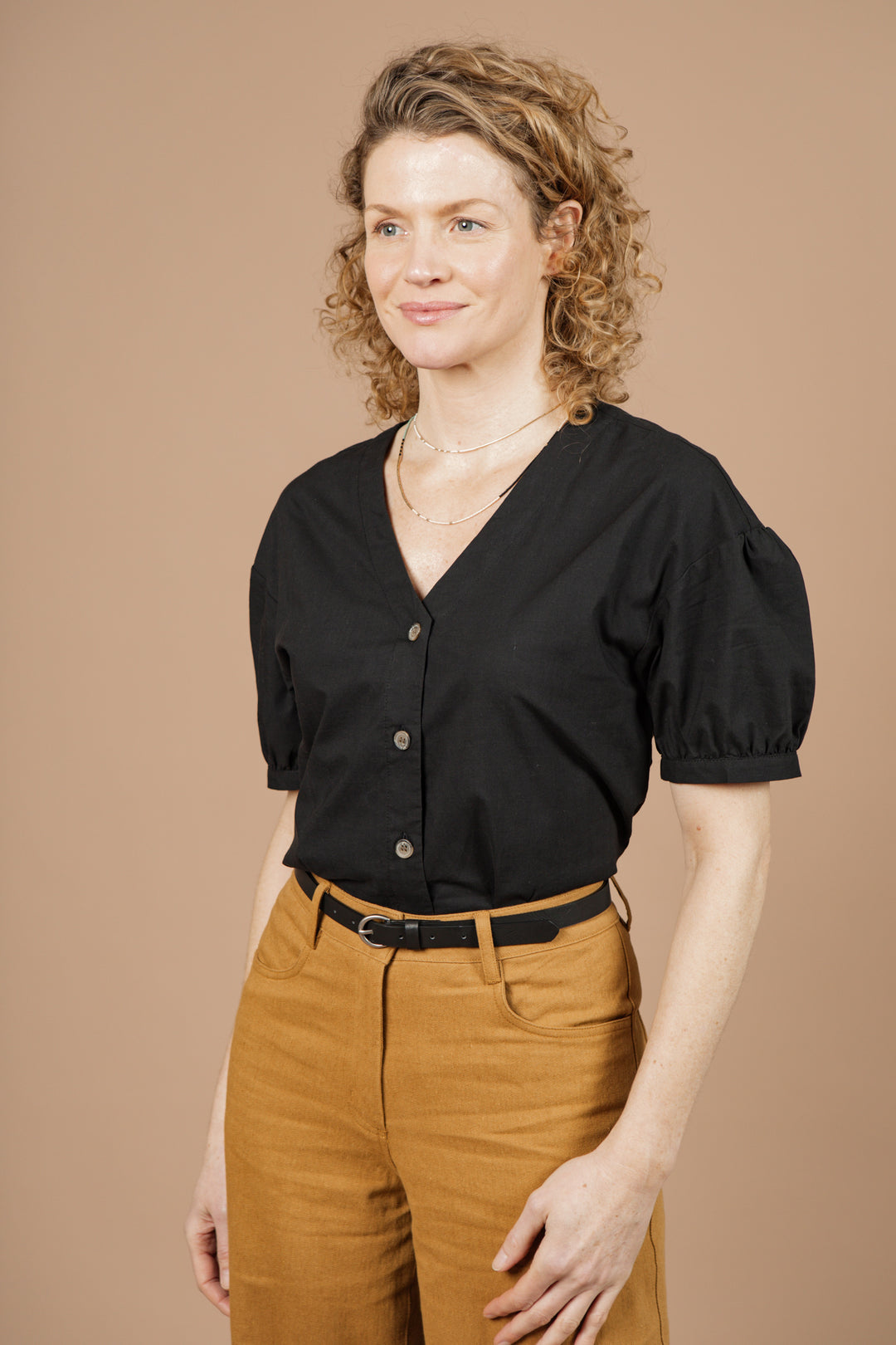 A woman with curly hair stands against a beige background, wearing the Charlotte Shirt in Black Poplin by Bridge & Burn. She pairs it with high-waisted tan pants and a black belt, smiling slightly as she looks to the side.