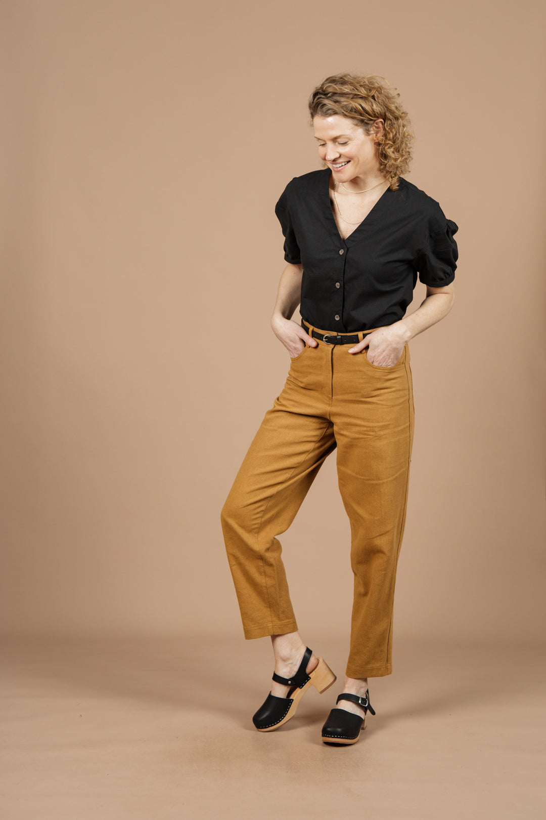A woman with curly hair wears the Bridge & Burn Charlotte Shirt in Black Poplin, high-waisted mustard pants, and black clogs. She stands against a beige background, smiling and looking down with one foot slightly raised.