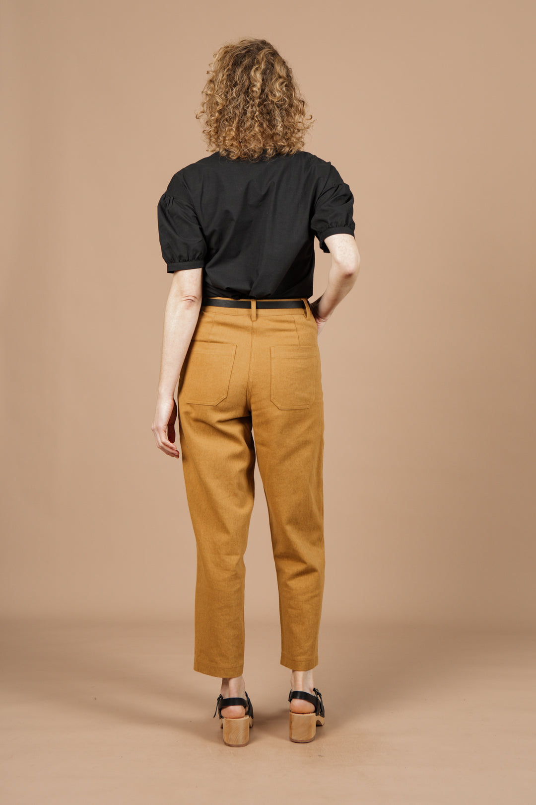 A person with curly hair, wearing the Bridge & Burn Charlotte Shirt in black poplin, tan high-waisted pants, and black sandals, stands facing away against a solid beige background.