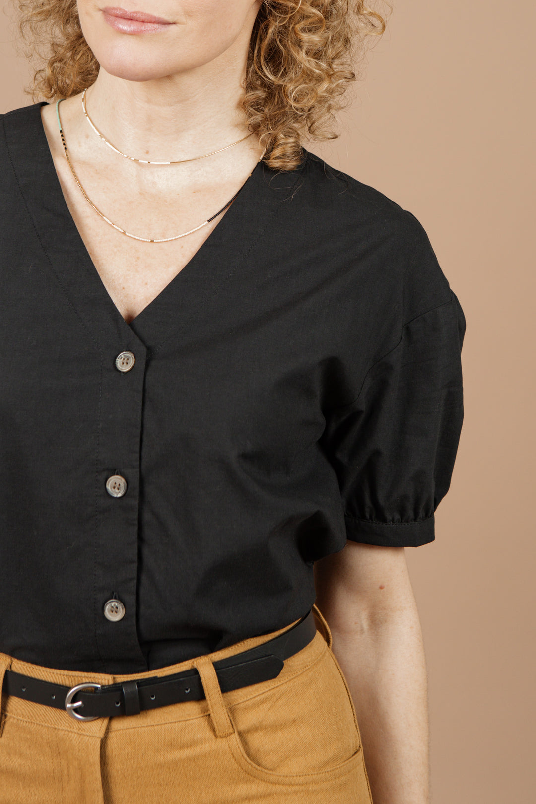 A woman with curly blonde hair wears the Bridge & Burn Charlotte Shirt in black poplin, featuring a V-neck and gathered sleeves, paired with high-waisted mustard trousers. She stands against a beige background, shown from the waist up.