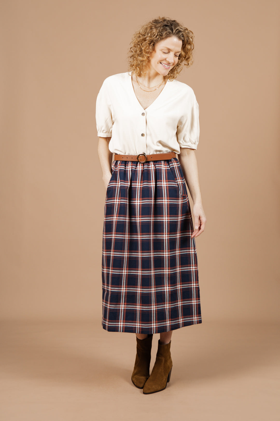 A woman with curly hair smiles against a beige background, wearing the Charlotte Shirt in cream poplin by Bridge & Burn, styled with a high-waisted navy plaid midi skirt and brown suede ankle boots.