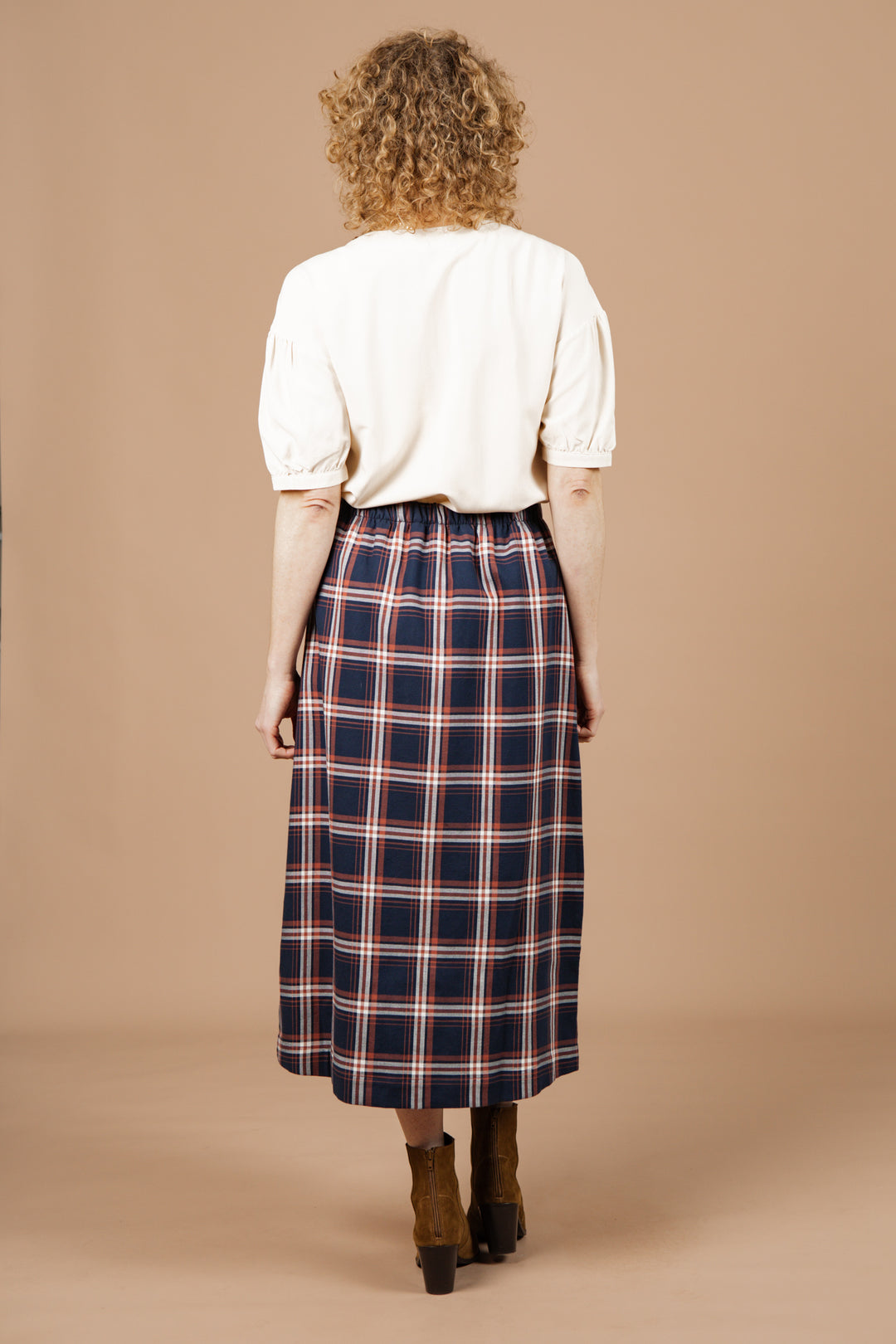 A person with curly hair, facing away, wears the Bridge & Burn Charlotte Shirt in cream poplin and a long blue, red, and white plaid skirt with brown ankle boots against a plain beige background.