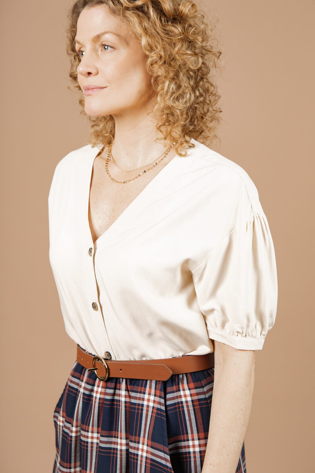 A woman with curly blonde hair wears the Bridge & Burn Charlotte Shirt in Cream Poplin paired with a brown belt and navy plaid skirt. She stands against a beige background, looking to the side.