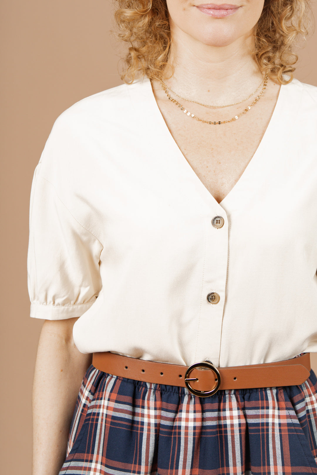 A person with curly hair wears the Bridge & Burn Charlotte Shirt in Cream Poplin, paired with a gold necklace, brown round-buckle belt, and a blue and red plaid skirt against a beige background.